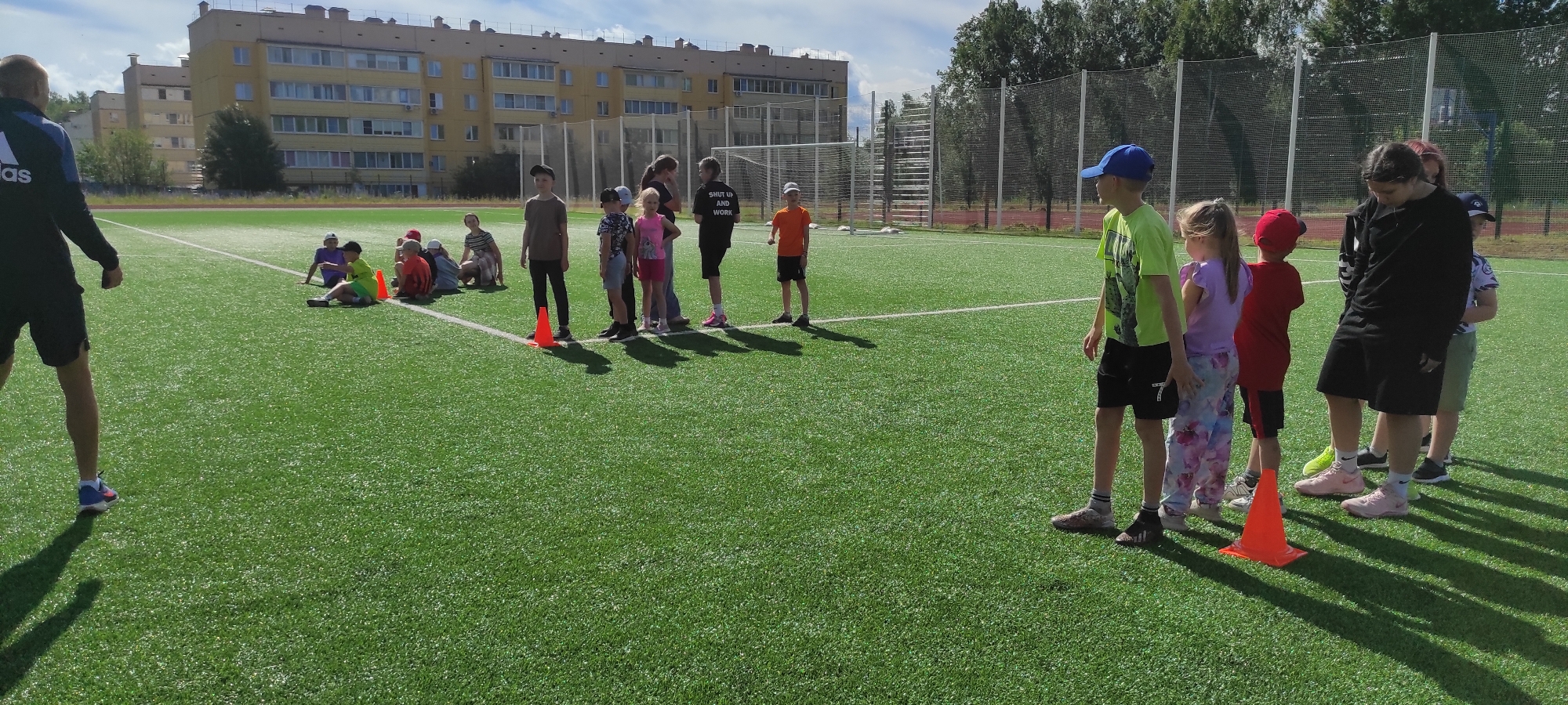 The image depicts a sunny outdoor scene on a green turf field, likely a sports ground. In the foreground, a person in dark athletic wear with a blue Adidas logo is partially visible on the left, facing away from the camera. A group of children is gathered on the field. Some are sitting in a line on the ground, while others are standing. There are orange cones placed on the field, suggesting some form of training or activity. In the background, a tall building with multiple floors and balconies is visible, along with trees and a sports net structure. The sky is partly cloudy. The overall environment suggests a public sports facility within the city of Noviy, Russia, being utilized for an outdoor group activity involving children.