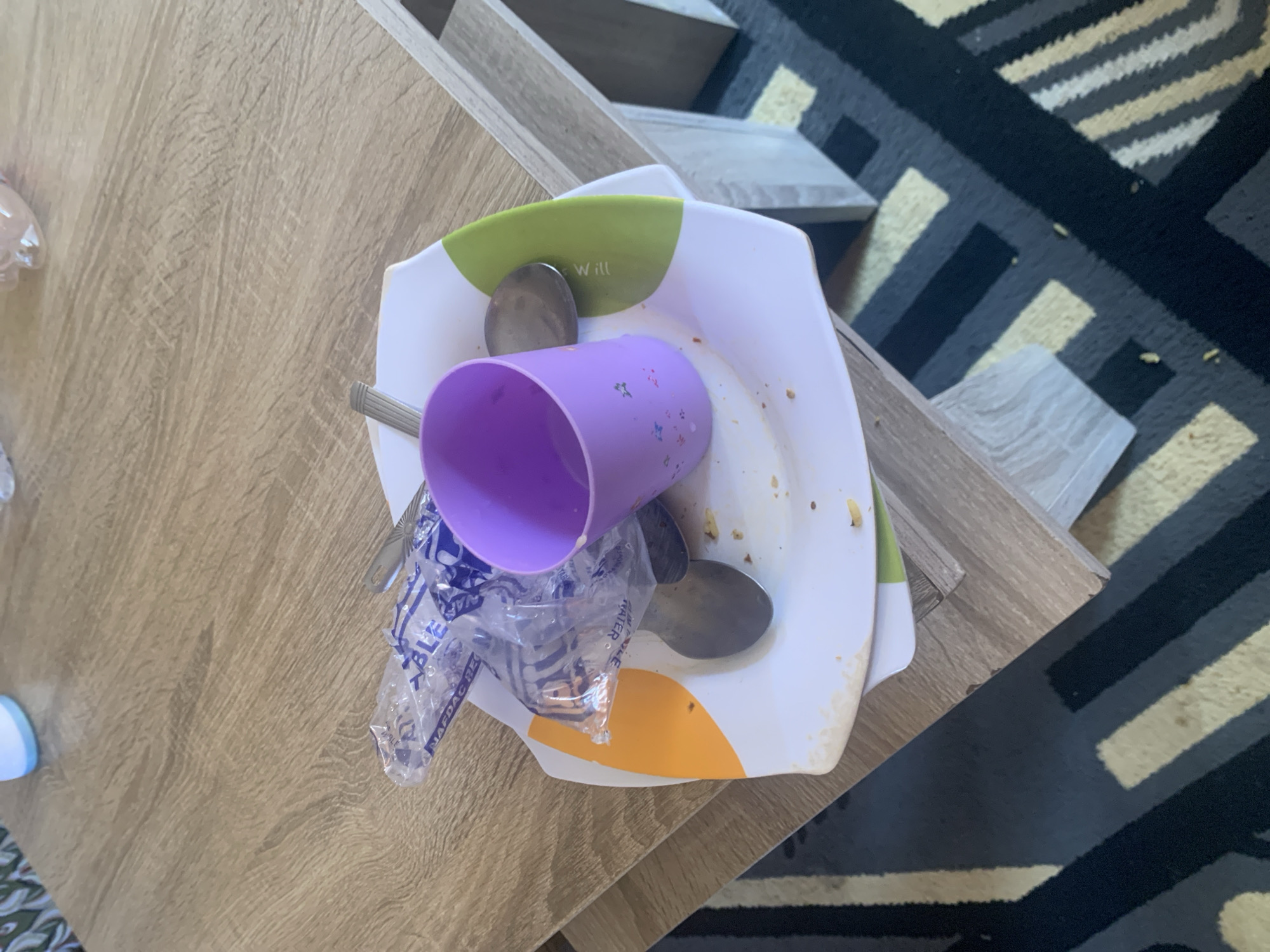 The image displays a dirty plate with a purple cup and cutlery on a wooden table. There is also a plastic wrapper with text visible. The plate appears to have food remnants on it. The background shows a carpet with a geometric pattern. The image is taken from a high angle. The location is identified as Gura Top, Nigeria. There are no people or specific actions depicted.