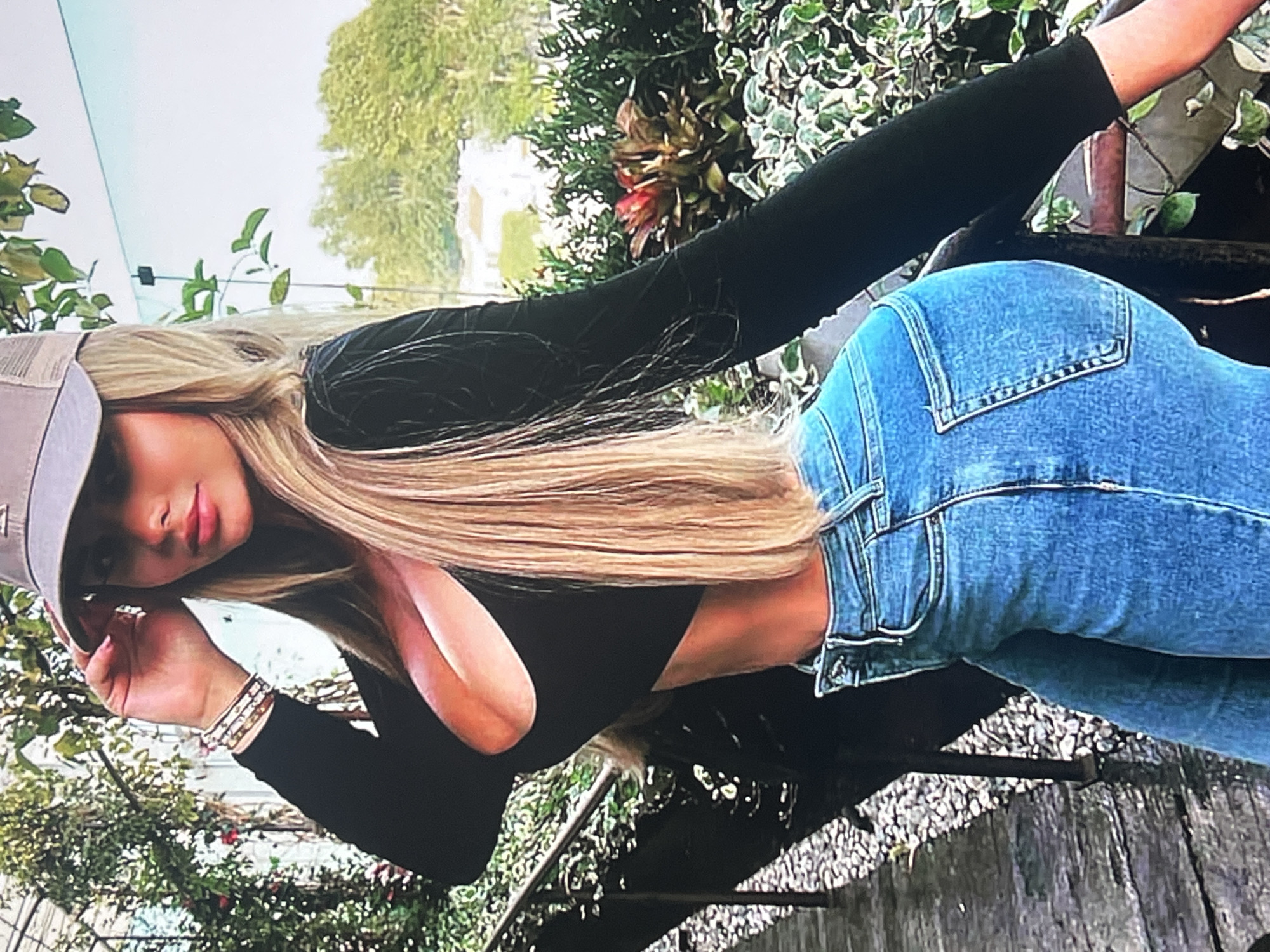A blonde woman wearing a black long-sleeved crop top, blue jeans, and a beige baseball cap is posing outdoors. She is standing on a walkway with gravel and wooden planks, with a metal railing on her right. Lush green foliage and some red plants are visible in the background, along with a clear sky and some distant trees. The woman is wearing bracelets on her left wrist.