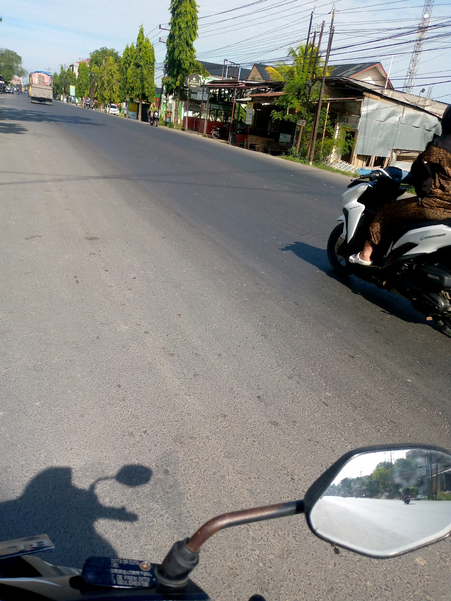 The image captures a view from a motorcycle on a paved road in Keudebayu, Indonesia. The road is lined with trees and buildings, suggesting an urban or semi-urban environment. In the foreground, the handlebars and a portion of the motorcycle's body are visible, along with a side mirror reflecting the road ahead. The side mirror shows other vehicles, including a motorcycle, on the road. Further down the road, a truck and several other motorcycles are seen. A person is riding a white scooter on the right side of the road, wearing a brown, patterned garment and white footwear. The sky is clear and blue, indicating daytime. The overall scene depicts everyday traffic and street life in the specified location.