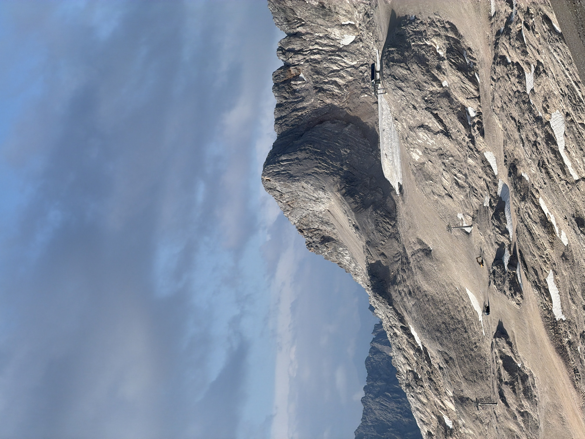 The image depicts a rugged, mountainous landscape under a cloudy sky. The terrain is characterized by rocky slopes, sparse patches of snow, and areas of exposed earth and gravel. In the foreground and midground, there are several man-made structures. These include ski lift towers, a small building or shelter on a plateau, and what appear to be construction vehicles, including an excavator and a utility vehicle. The overall impression is of a high-altitude, possibly alpine environment, perhaps a ski resort area undergoing maintenance or preparation. The specific location within Garmisch-Partenkirchen, Germany, is likely a high mountain area accessible by cable car or ski lift, such as the Zugspitze or a related ski area, given the infrastructure and terrain. There are no people visible in the image.