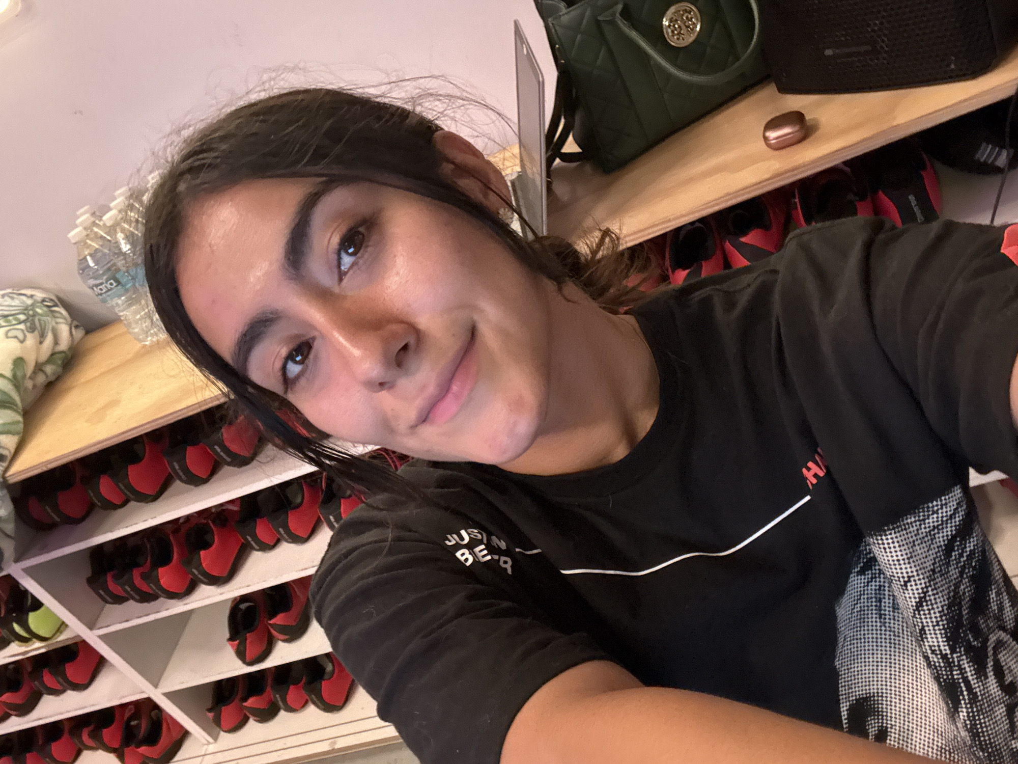 A person with long, dark hair pulled back and brown eyes is smiling at the camera. They are wearing a black T-shirt with white accents and are positioned in front of shelves filled with red and black shoes. Above the shelves are a wooden surface with a green purse, a black speaker, and a copper-colored object. Behind the person on the left are stacked water bottles and a patterned cushion on a wooden surface. The location is likely a climbing gym shoe rental area. It is not possible to confirm the location in the city of Puebla, Mexico, from the image.