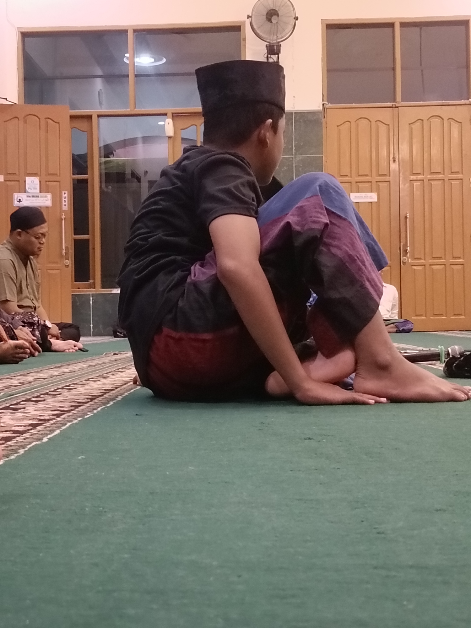 The image captures an interior scene, likely a prayer room or a community gathering space, given the presence of multiple individuals in religious attire and the carpeted floor. In the foreground, a young boy wearing a black peci (a brimless cap commonly worn by Muslim men and boys) and a black t-shirt is seated on a green carpeted floor. His legs are bent, and he is wearing patterned trousers in shades of blue and maroon. He is looking towards the right side of the frame. To the left, an adult male, also wearing a peci and a light brown shirt, is seated on the same carpet, facing left. His hands are resting on his lap. Behind them, there are wooden doors with ornate panels and windows. A ceiling fan is visible in the upper portion of the image. The overall atmosphere suggests a serene and communal setting. The visual clues, such as the prayer room setup and the attire of the individuals, are common in mosques or religious centers found throughout Kediri City, Indonesia.
