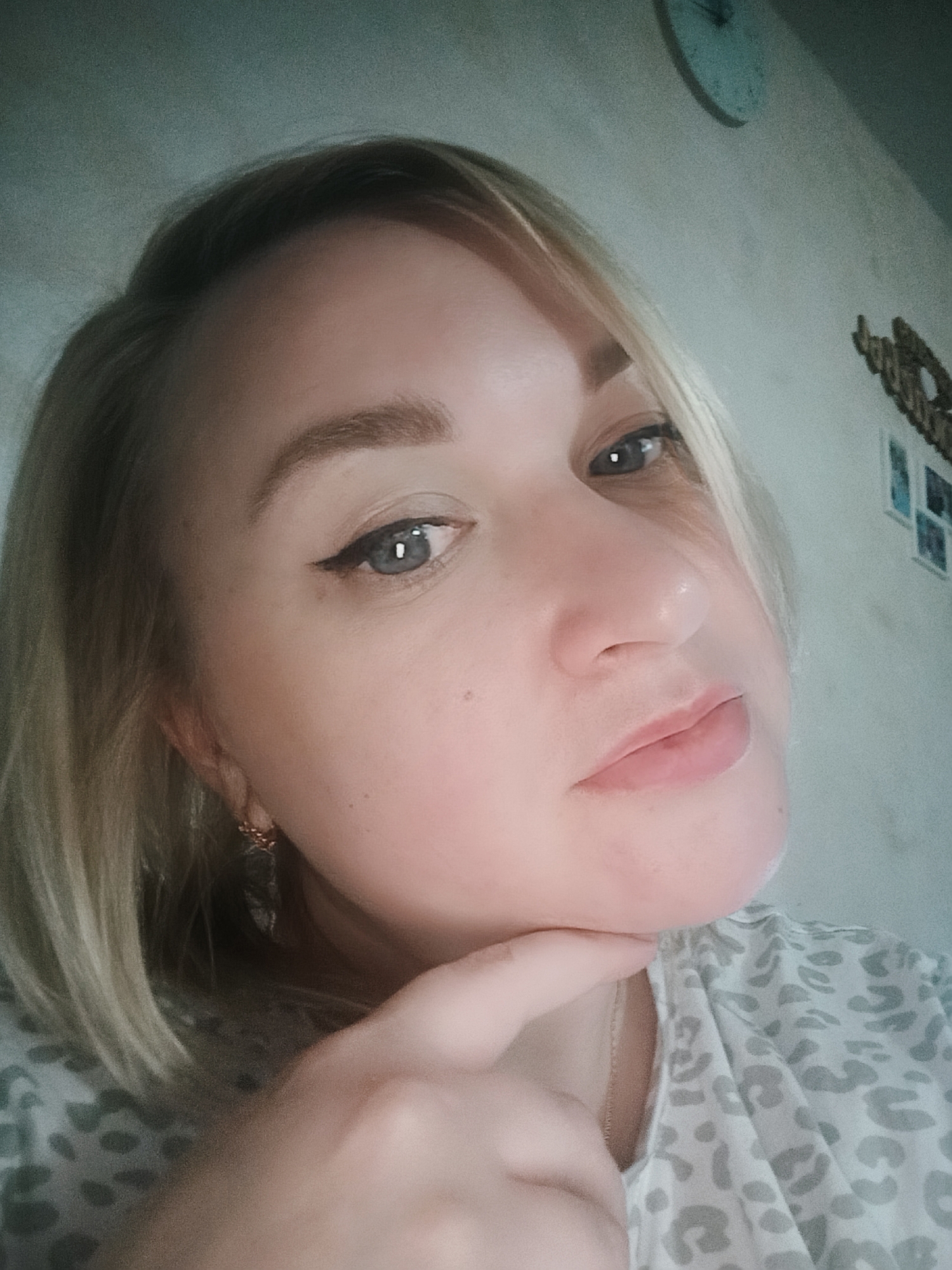 A close-up, slightly angled shot of a fair-skinned woman with light blonde hair. She has light blue eyes with black eyeliner. Her left index finger is touching her chin. She is wearing a light-colored patterned shirt, and a small gold earring is visible in her left ear. In the background, a wall is visible with a clock and some decorative items or pictures. The location is not identified.