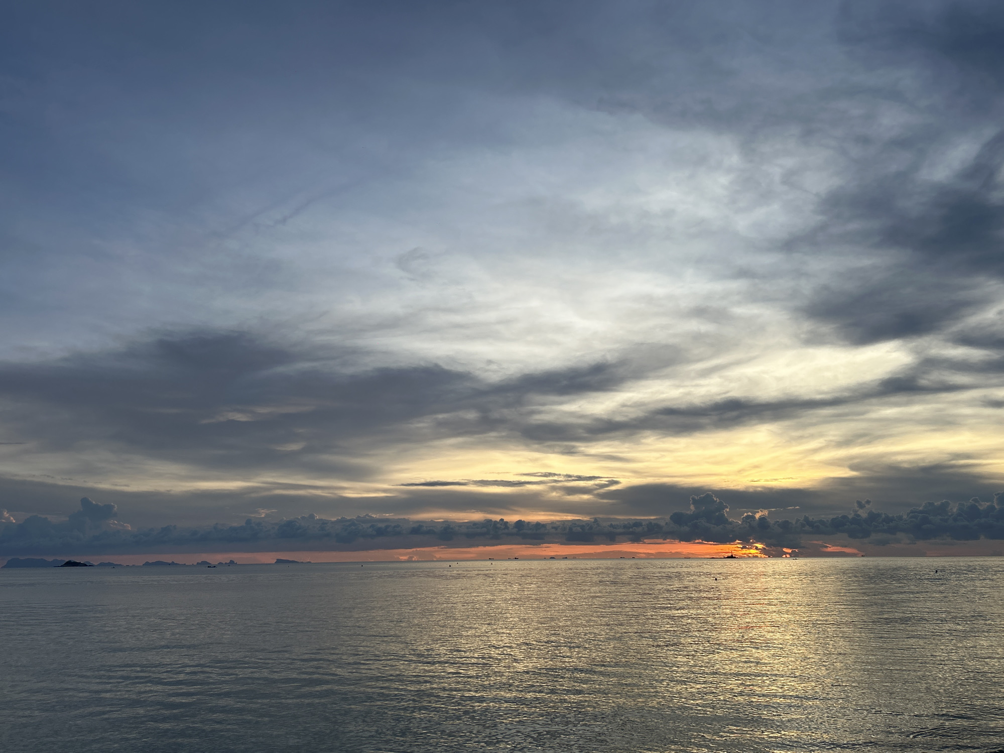 The image shows a seascape under a cloudy sky, likely taken near Baan Sri Thanu, Thailand. The sky is filled with grey and white clouds, with hints of orange and yellow light near the horizon, suggesting either sunrise or sunset. The water is relatively calm with reflections of the sky and light. In the distance, there are land masses, possibly islands or peninsulas, partially obscured by the horizon's haze. There are some scattered objects on the water which may be boats, birds or people swimming in the water. The overall scene is serene and atmospheric.