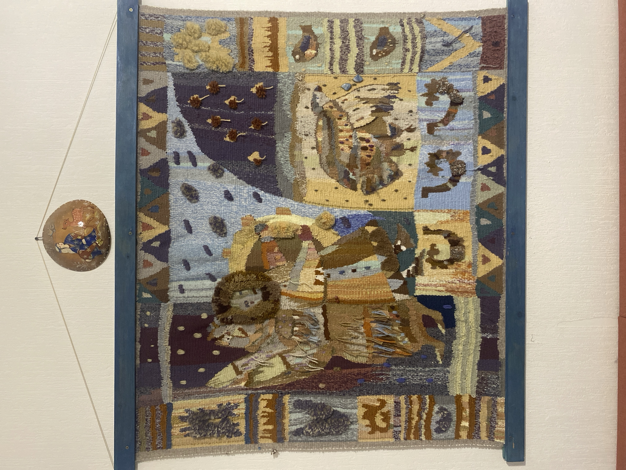 A framed tapestry hangs on a white wall with a light peach accent wall section on the right side of the frame. To the left of the tapestry, attached to the frame by a string, is an Asian-style lacquered bowl. The tapestry features a scene with earth-toned colors, depicting what appears to be a figure riding an animal. The style is abstract and textural, with various patterns and materials used in the weaving. The frame of the tapestry is dark blue. The location is an interior space; there is no information about the city of Yekaterinburg, Russia.