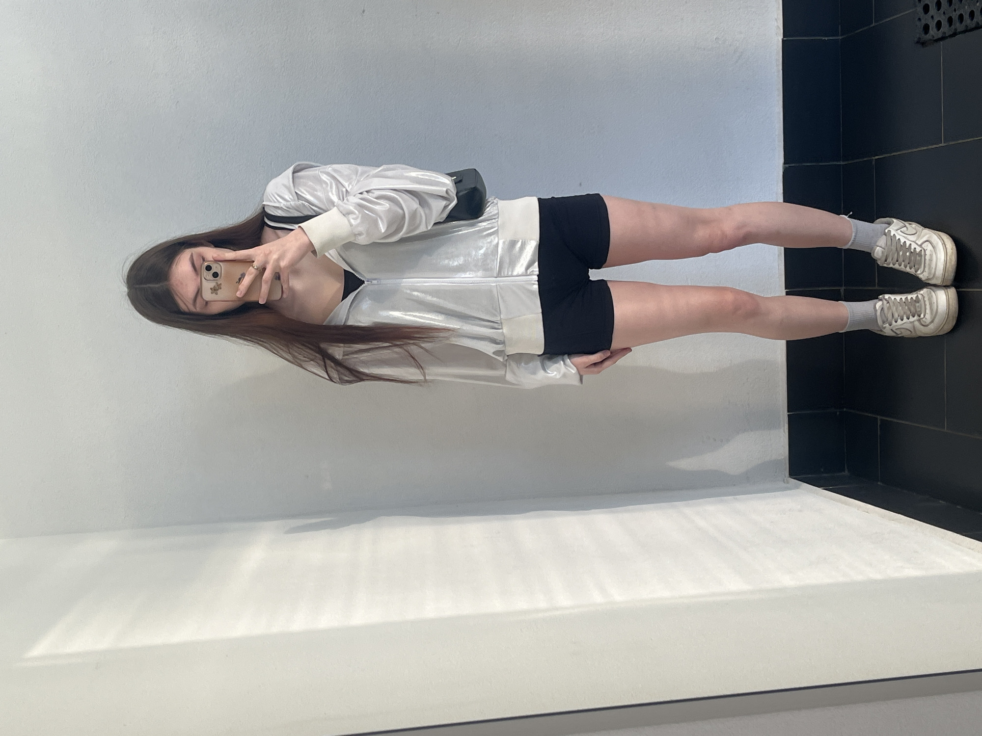 The image depicts a young woman taking a mirror selfie indoors in Saint Petersburg, Russia. She has long, straight, dark brown hair, and her face is obscured by a light-colored smartphone with a patterned case, which she holds with her right hand. A gold-colored ring is visible on her right ring finger. She wears a shimmering silver full-zip hoodie with off-white ribbed cuffs and waistband, over a black sports bra or crop top. Her lower body is clad in black form-fitting shorts, grey ankle socks, and worn white athletic sneakers. A small black shoulder bag hangs over her left shoulder. The background features plain white walls and a black tiled floor. Diagonal patterns of light and shadow are cast on the left wall, indicating an external light source. A dark rectangular mat is partially visible on the floor in the lower right corner.