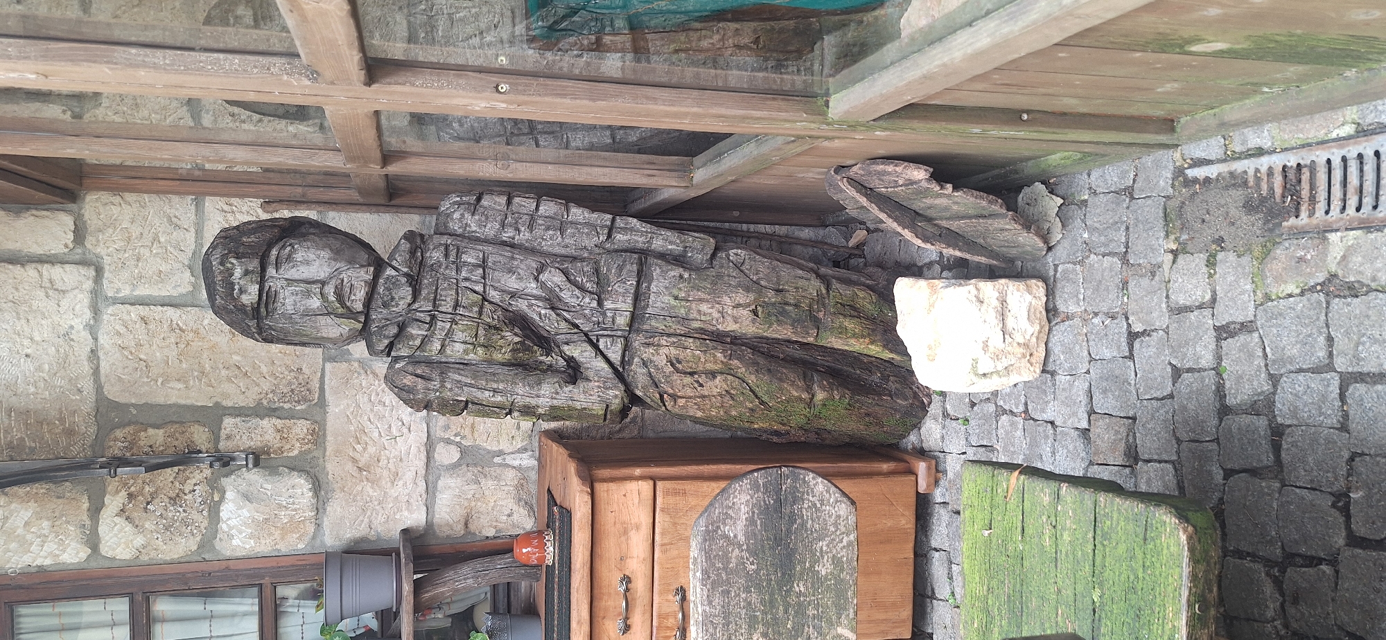 The image shows a wooden sculpture of a figure standing in an outdoor area paved with cobblestones. The sculpture is carved from wood and appears weathered, with some moss growing on it. It is dressed in what looks like armor or a patterned garment. To the left of the sculpture is a wooden chest of drawers with metal handles and a round wooden tabletop placed on it. In the background, there is a stone wall. To the right of the sculpture is a wooden structure with glass panes, resembling a door or window. There is a white stone block and a broken piece of wood lying on the ground near the sculpture. A green wooden bench is visible in the lower left corner of the image, and a metal drain grate is present in the lower right. The scene is likely in a courtyard or garden area. The location is identified as Kalimantsi, Bulgaria.