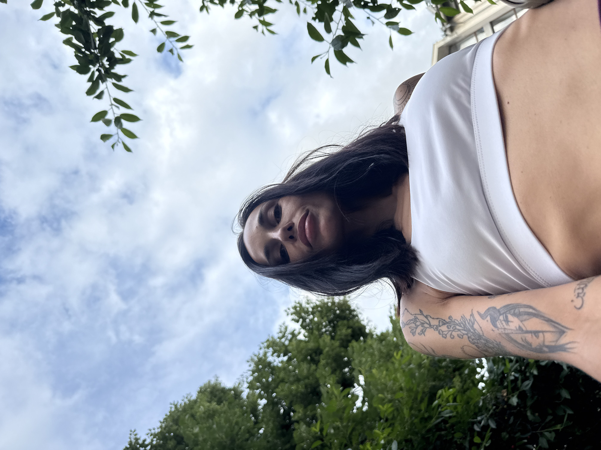 An upward-angled shot captures a woman with dark hair and fair skin, looking directly down at the camera. She wears a white top or swimsuit, with her face prominently in the upper central part of the frame. Her expression is calm, with her lips slightly parted. Visible on her right arm are several tattoos: a detailed floral or botanical design extends along her forearm, and a small, possibly numeric character like a '3' is inked closer to her shoulder.
The background is dominated by a bright, cloudy sky featuring scattered white clouds. Green tree foliage, with distinct leaves and branches, frames the top and bottom sections of the image, suggesting an outdoor, natural environment such as a park or garden. In the upper right corner, a light-colored building with window reflections is faintly visible, indicating an urban or suburban setting beyond the immediate greenery. The natural lighting suggests it is daytime, likely late morning or early afternoon, under a partially overcast sky. The overall scene appears peaceful, set in Pleven, Bulgaria.