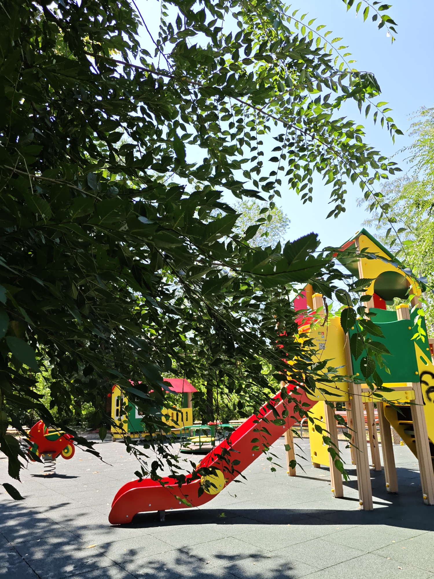 The image depicts a children's playground in Rostov-on-Don, Russia. The playground features a colorful slide, a spring rider shaped like a motorcycle, and a playhouse structure. Lush green trees with visible leaves partially obscure the view of the playground equipment and the clear blue sky. The ground surface appears to be a dark, padded material, and shadows from the trees are cast across it. The scene is set outdoors on a sunny day, with the vegetation indicating a warm season. There are no people visible in the image.