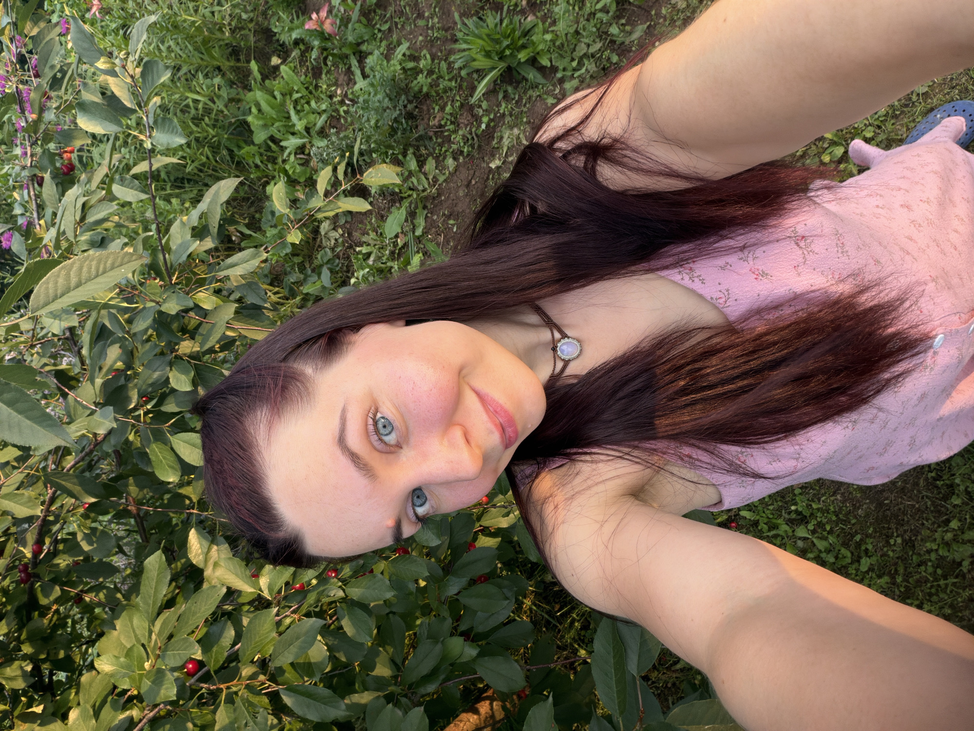 A young woman with long, dark hair and blue eyes is taking a selfie outdoors. She is wearing a light pink dress with a floral pattern and a necklace with a light-colored pendant. She is standing in front of a tree with green leaves and red cherries. The background shows lush green foliage and some wildflowers. The lighting suggests it is late afternoon or early evening, with warm sunlight casting a glow on her arm. The location is identified as the city of Фоки, Russia.