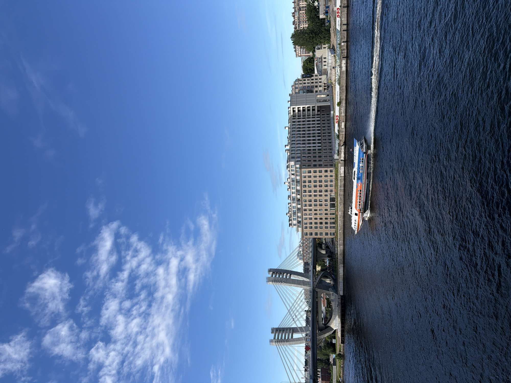 The image shows a hydrofoil boat moving across a body of dark blue water, leaving a white wake behind it. In the background, a modern cable-stayed bridge with two distinctive pylons spans the water. To the right of the bridge, a multi-story, light-colored apartment building with numerous windows is visible, along with other buildings and trees further in the distance. The sky is bright blue with scattered white clouds. This scene is located on the Neva River in Saint Petersburg, Russia, with the Western High-Speed Diameter bridge and apartment complexes characteristic of the city's modern waterfront development.