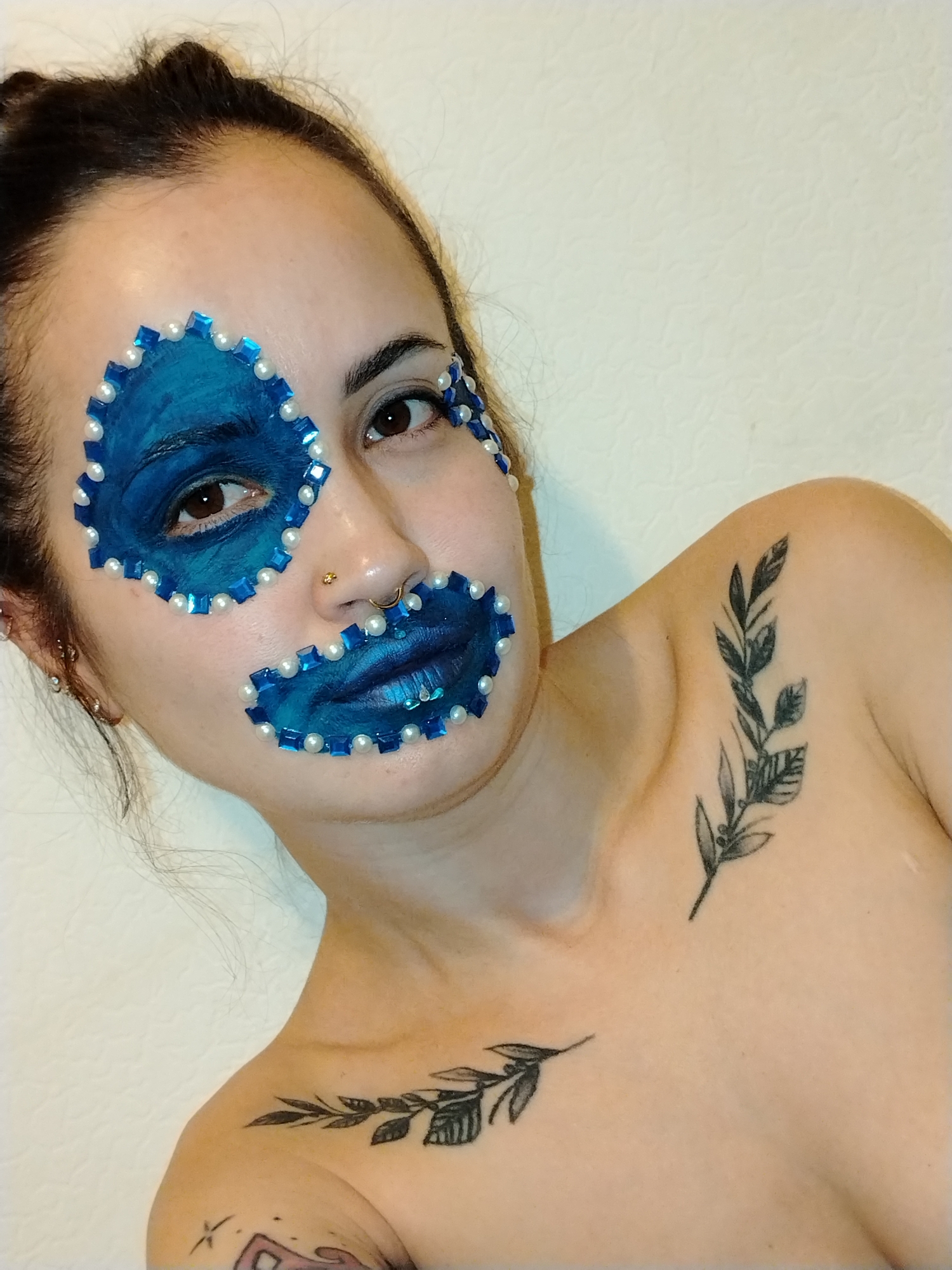 A close-up studio portrait shows a light-skinned woman with her dark hair pulled back. She has blue face paint around her right eye and mouth, decorated with small white pearls and blue square gems. A nose ring is visible, and she has tattoos of leafy branches on her chest and collarbone. The background is a plain, light-colored wall. The city of Noviy, Russia is not discernible from the image.