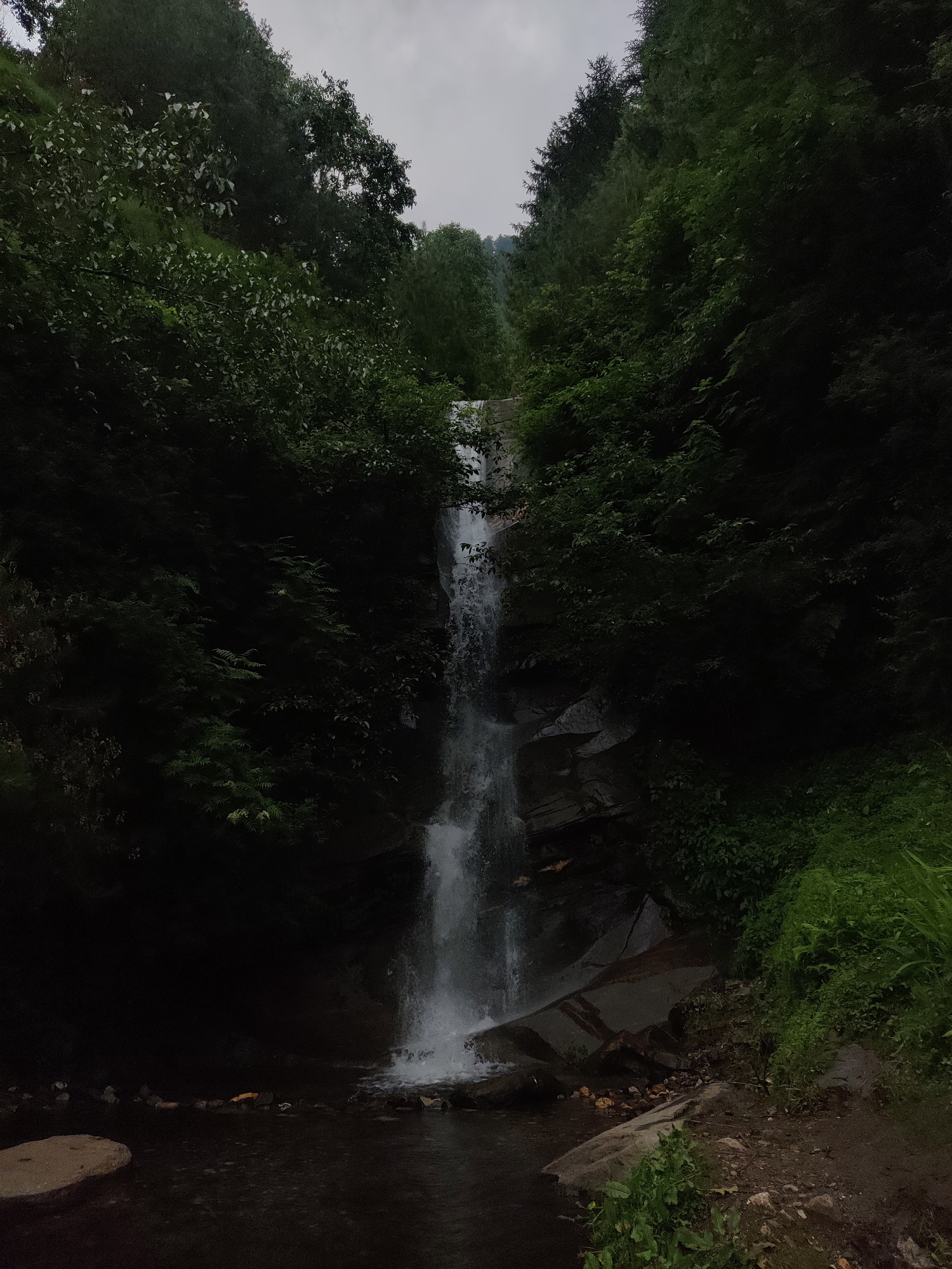 The image depicts a tall, narrow waterfall cascading down a rocky, heavily wooded mountainside. The water is white and frothy as it tumbles over dark, wet rocks. Lush green foliage, including dense trees and undergrowth, surrounds the waterfall, creating a secluded and natural setting. The sky above is overcast, casting a muted light on the scene. The foreground shows a dark, still pool of water at the base of the falls, with some rocks and a sandy bank visible. The overall impression is one of a wild and somewhat somber natural landscape, possibly located in a mountainous region like the one surrounding Vashisht, India, known for its natural beauty and waterfalls. There are no people or man-made structures immediately apparent in the image.