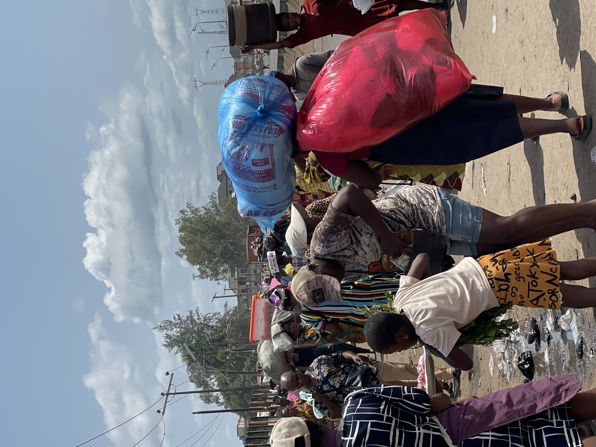 The image captures a bustling market scene in Onitsha, Nigeria. In the foreground, vendors are seen interacting with customers amidst scattered litter on the ground. One woman carries a large red bag and a blue bag on her head. The blue bag has text reading "GOOD MEN'S POLY." A man walks behind her carrying a large metal container on his head. In the background, buildings, trees, and power lines punctuate the cityscape under a partly cloudy sky. The scene suggests a typical day of commerce and activity in the city.