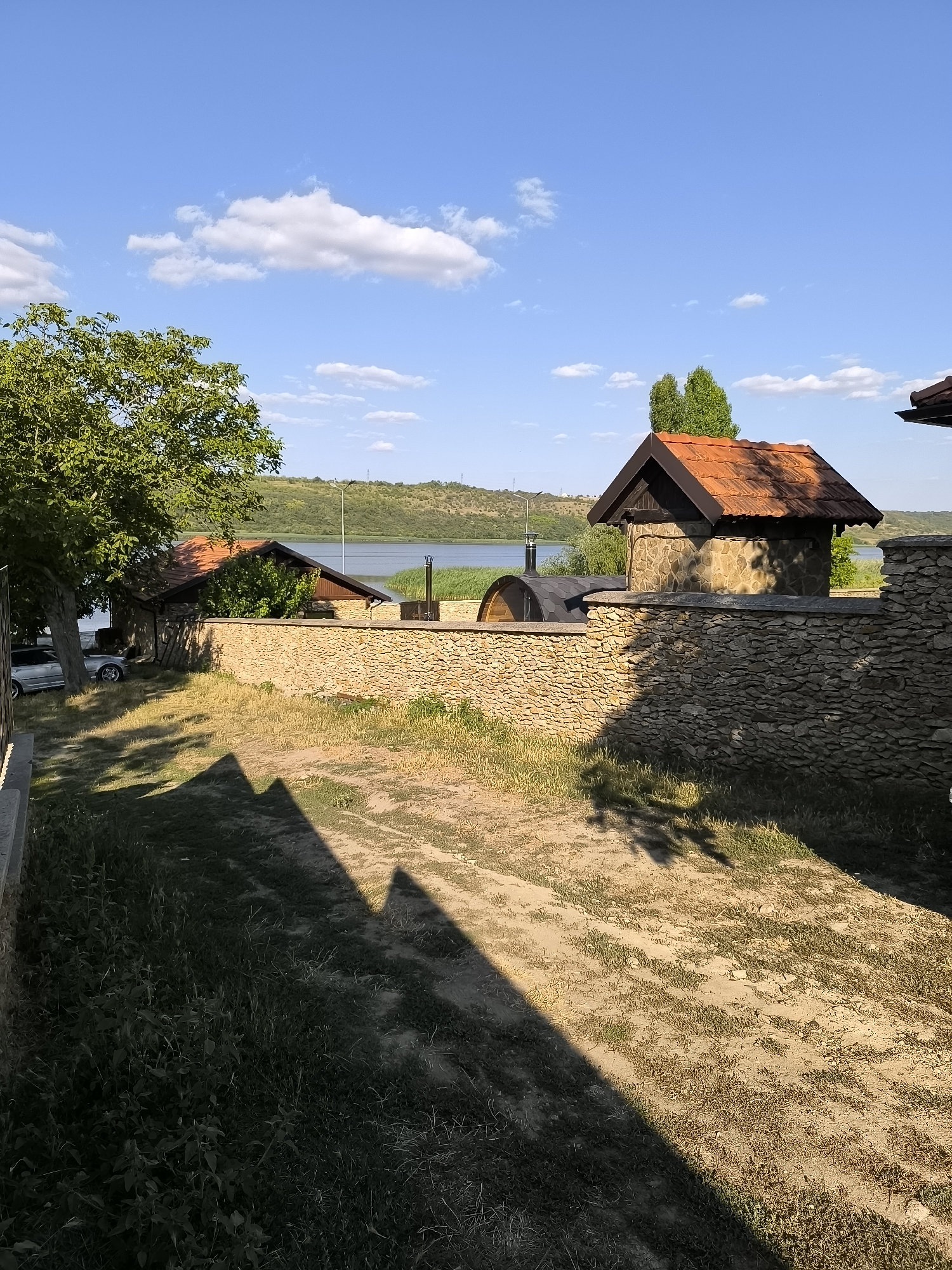 The image depicts a sunny outdoor scene in Jora de Mijloc, Moldova, featuring a rustic stone-walled compound next to a body of water, likely the Jora de Mijloc Reservoir. The compound contains two small buildings with reddish-brown tiled roofs, constructed with rough-hewn stones. One building is larger, resembling a small house or outbuilding, while the other is a smaller, more enclosed structure with a dark roof. A curved, barrel-shaped structure, possibly a sauna or storage, is visible behind the stone wall.
The stone wall itself is a prominent feature, built from irregularly shaped stones and stretching across the foreground and midground of the image. Beyond the wall, a wide, calm body of water is visible, bordered by lush green hills in the distance. The sky is a vibrant blue with scattered white cumulus clouds.
In the left of the image, a large, leafy green tree provides shade, and a silver car is parked near the building. The ground in the foreground consists of dry grass and dirt, with distinct shadows cast from an unseen structure or trees, creating a geometric pattern on the ground. The overall impression is one of a peaceful, rural setting, possibly a vacation or recreational area, with a focus on natural materials and integration with the landscape. There are no people visible in the image.