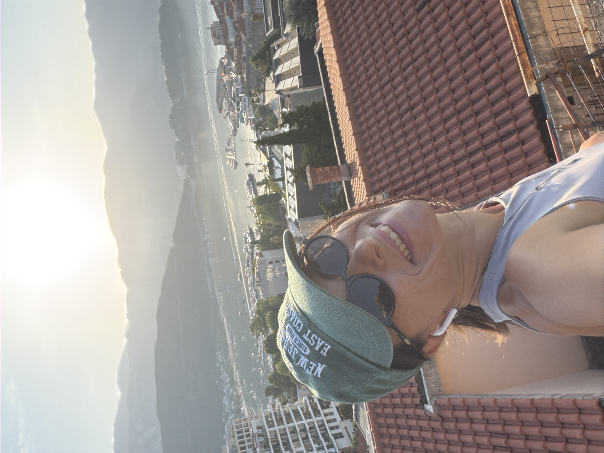 In Budva, Montenegro, a woman stands on a terracotta tiled rooftop, looking up and smiling at the camera. She is wearing a green baseball cap that reads "NEW JERSEY SINCE 1985 EAST COAST", large black sunglasses, and a light blue tank top. She has earbuds in her ears and a necklace with a pendant. Behind her, the scene unfolds with a picturesque view of the Budva harbor, dotted with numerous boats and yachts. The coastline is lined with buildings, ranging from modern high-rises to traditional houses with red roofs. In the distance, misty mountains form a dramatic backdrop against the bright, hazy sky, suggesting either sunrise or sunset. The overall ambiance is one of leisure and scenic beauty.