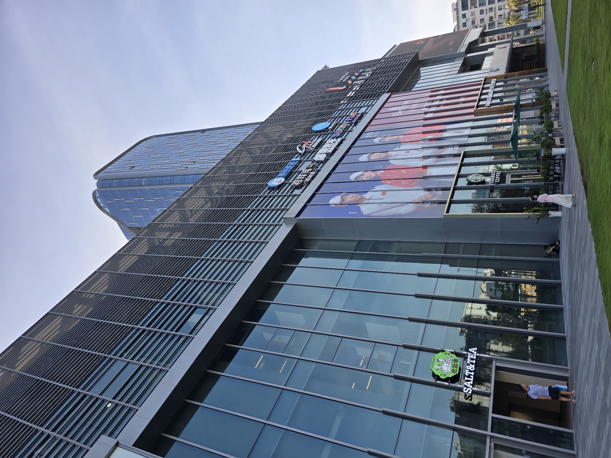 In Tianya District, China, a modern, multi-story commercial building with extensive glass facades and metal louvers stands under a clear sky. The building features several large advertisements, including one prominently displaying four individuals in athletic attire and another with the "STARBUCKS COFFEE" logo. A sign for "SALT&TEA" is visible on the lower level. People are present, including a man in a striped shirt standing near the entrance of "SALT&TEA," a woman in a pink dress walking on the paved area, and individuals seated at tables outside the Starbucks. Green grass and palm trees are part of the surrounding landscape. The architecture is contemporary, with reflective glass and sharp geometric lines.