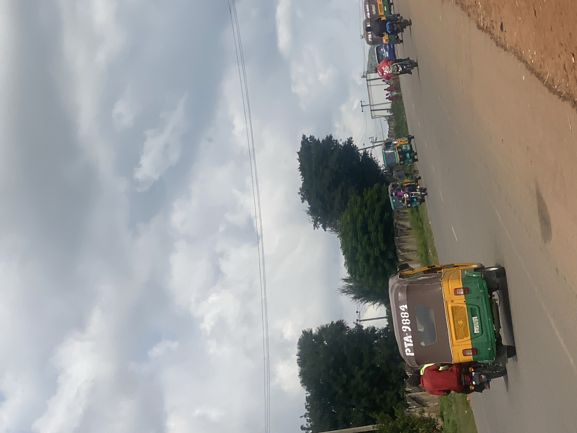 The image depicts a street scene in Rafin Par, Nigeria, under a cloudy sky. The foreground is dominated by a yellow and green auto-rickshaw with the registration number PTA-9884, driven by a man in a red shirt. Behind him, another auto-rickshaw is partially visible. The road stretches into the distance, where several other auto-rickshaws and motorcycles are seen. On the right side of the road, there are buildings and people, with some of the auto-rickshaws displaying "PTA 2815" and "PTA 2818" on their roofs. Trees line the left side of the road, and a concrete wall is visible behind them. Utility poles and wires are present in the background.