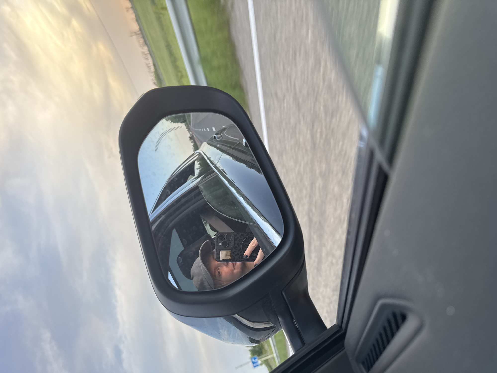 The image shows a side mirror of a car reflecting a person wearing a cap and holding a phone. The background visible through the mirror suggests the car is moving on a road with green fields on the sides. The road is divided by a white line. The driver's reflection shows that they are in a car, and the rest of the scene seen in the mirror, including the sign, indicates the location may be in Fastovetskaya, Russia.