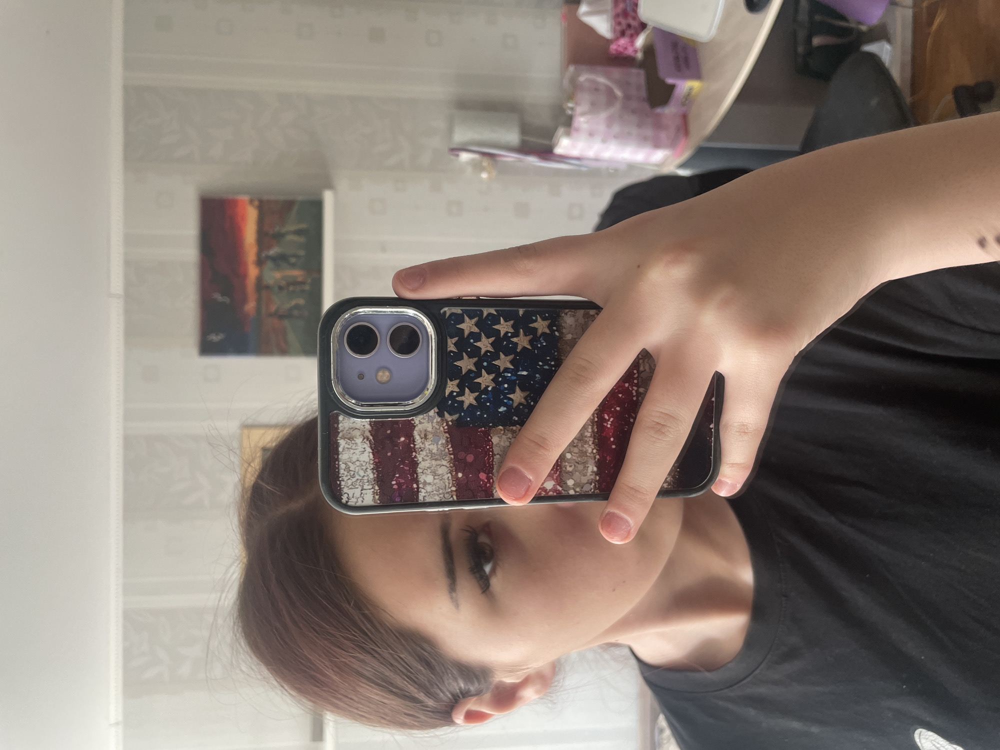 This close-up mirror selfie captures a person, likely a young female, holding a smartphone with a distinctive case. The image is rotated 90 degrees clockwise. The person wears a black t-shirt, and her right hand holds the phone, obscuring much of her face except for her right eye, ear, and a portion of her reddish-brown hair. Her expression appears neutral, focused on the act of taking the photo.
The smartphone, which features a dual-camera system, is encased in a glittery American flag design, showcasing red and white stripes and a blue field adorned with stars.
The reflection in the mirror reveals an indoor setting, consistent with a home environment in Sinyavino, Russia. Behind the person, a light-colored wall or wallpaper with a subtle patterned texture is visible. A small framed picture or abstract painting with deep red, orange, and darker hues hangs on this wall. Various other items, including what appears to be a tissue box, are reflected on a surface behind the person, suggesting a desk or shelf. The lighting is soft and diffused, indicating daytime. No visible text is present in the image.