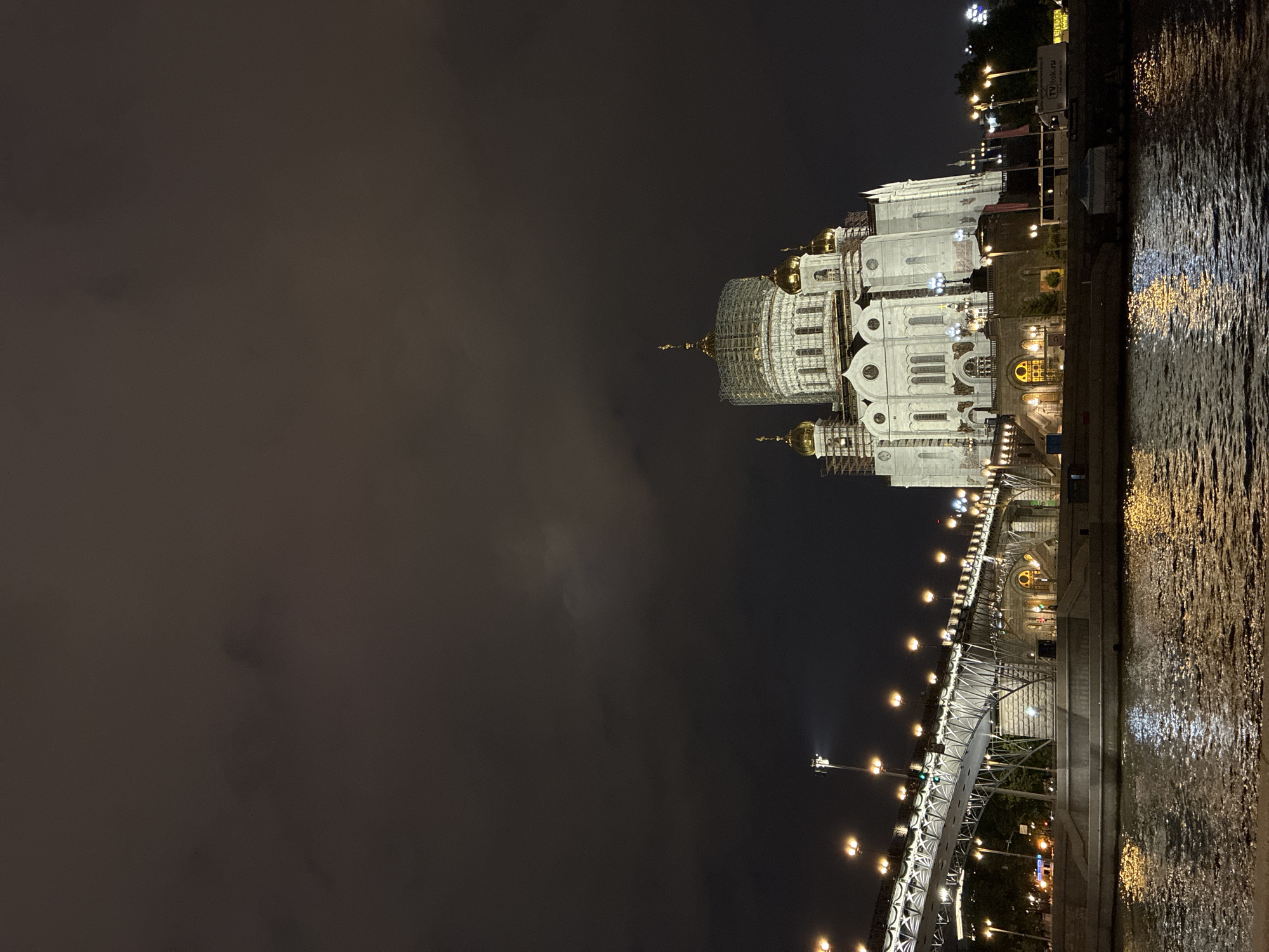 The image depicts the Cathedral of Christ the Savior in Moscow, Russia, at night. The cathedral, illuminated with white and yellow lights, is the dominant feature on the right side of the frame. Its golden domes are also highlighted, creating a striking visual against the dark sky. The Moscow River is visible along the right edge, with reflections of the lights shimmering on the water's surface. A bridge, also lit with numerous lights, spans the river and leads towards the cathedral. The left side of the image is filled with a dark, cloudy sky.