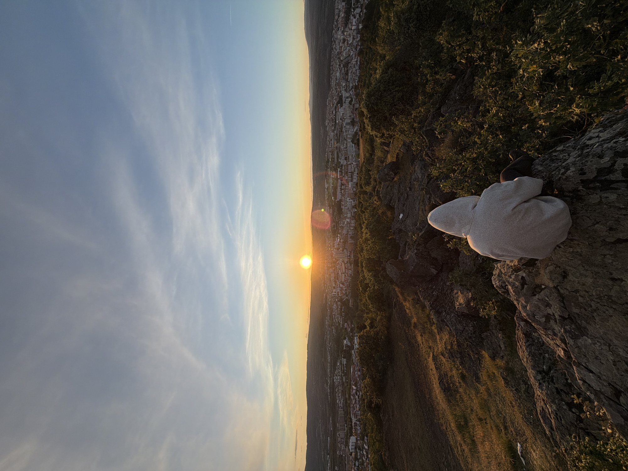 The image shows a person in a light gray hoodie and dark pants sitting on a rocky outcrop overlooking a city at sunset. The sky transitions from light blue with wispy clouds to a golden yellow near the horizon where the sun is setting. The city below appears to be Bragança, Portugal, based on the urban layout and surrounding terrain. The ground near the rocky outcrop is covered with sparse vegetation. The image is oriented vertically, with the sky on the left and the ground sloping down to the city on the right.