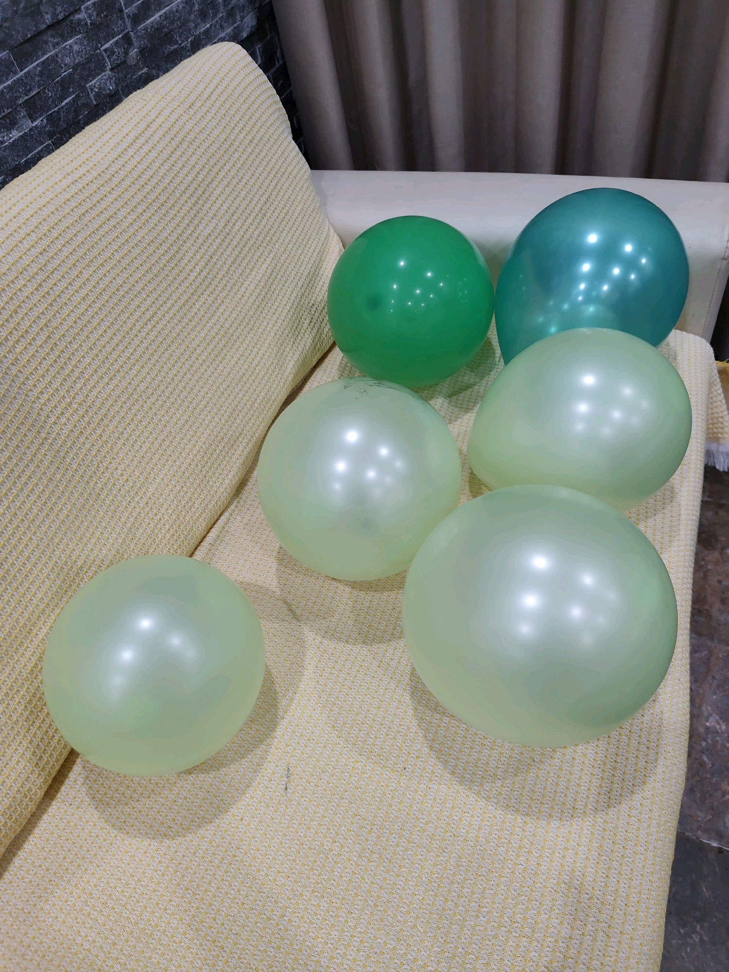 A group of six balloons is arranged on a couch. Two of the balloons are a bright green color, and the other four are a pale, translucent green. They are all inflated and appear to be made of latex. The couch has a textured, light yellow fabric. In the background, a dark stone wall is visible on the left, and draped curtains in a neutral color are on the right.