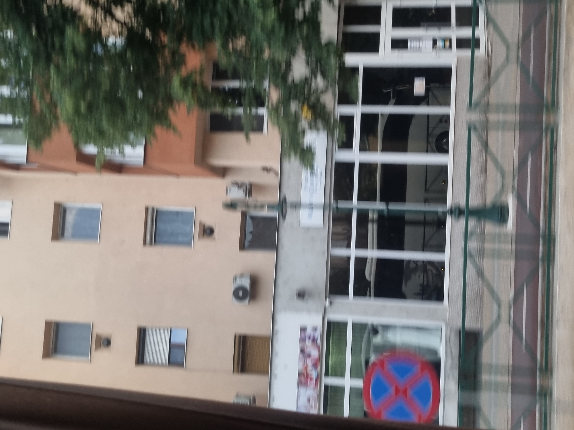 The image depicts a street view in Salonta, Romania, focusing on the facade of a building. The building has multiple floors with a light-colored exterior. Several windows are visible, some with blinds or shutters. There are also air conditioning units mounted on the exterior walls.
The ground floor of the building appears to house commercial establishments. A large storefront with multiple windows is visible, displaying items that suggest retail or service businesses. A red and blue circular sign with a red cross is positioned in front of one of the windows. A green metal fence with a decorative pattern runs along the sidewalk in the foreground. A street lamp or pole is also present. Trees with green foliage are seen in the background, partially obscuring the upper floors of the building and an adjacent building with an orange-brown facade. The overall impression is of an urban setting, possibly a main street or commercial area.