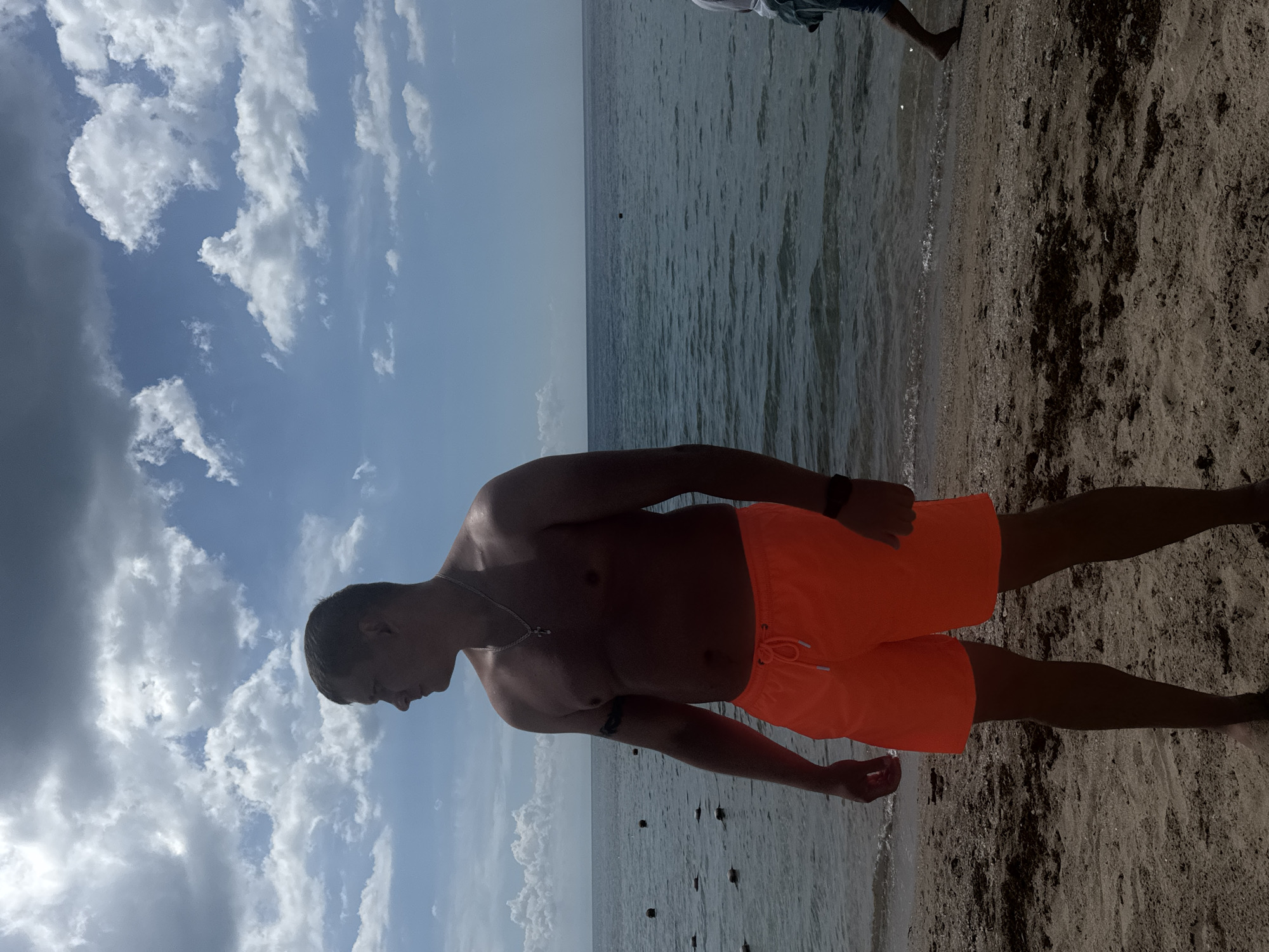 A man in orange swim trunks stands on a sandy beach beside the water. The location appears to be Yevpatoriya, Ukraine. The sky above features blue with puffy white clouds. In the water, there are dark floating objects.