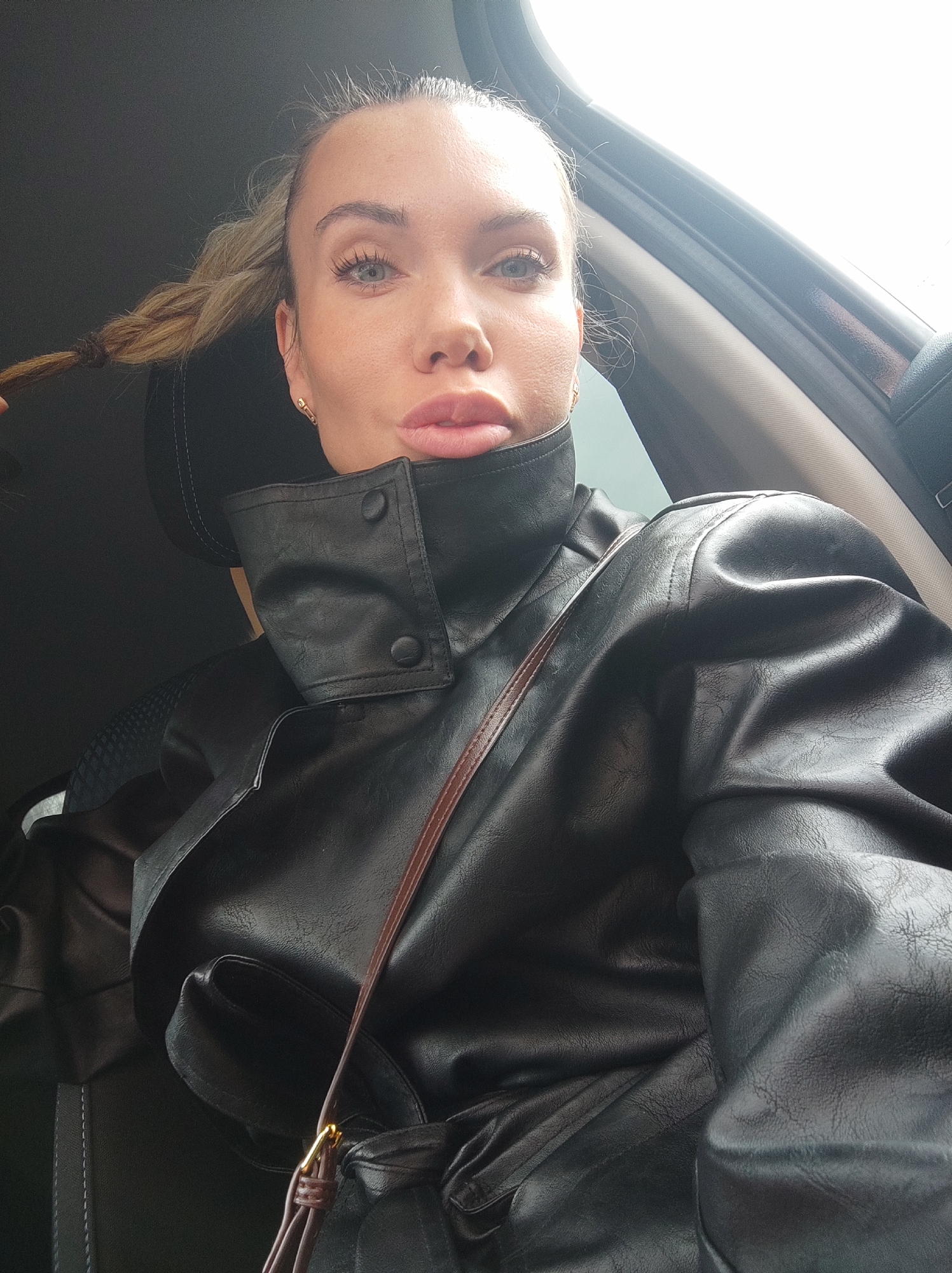 A woman is seated in a vehicle. She is fair-skinned with blonde hair styled in a braid. She wears a black faux leather trench coat. The environment suggests the interior of a car, likely in Votkinsk, Russia. The specifics of her actions or interactions are not discernible from the image.
