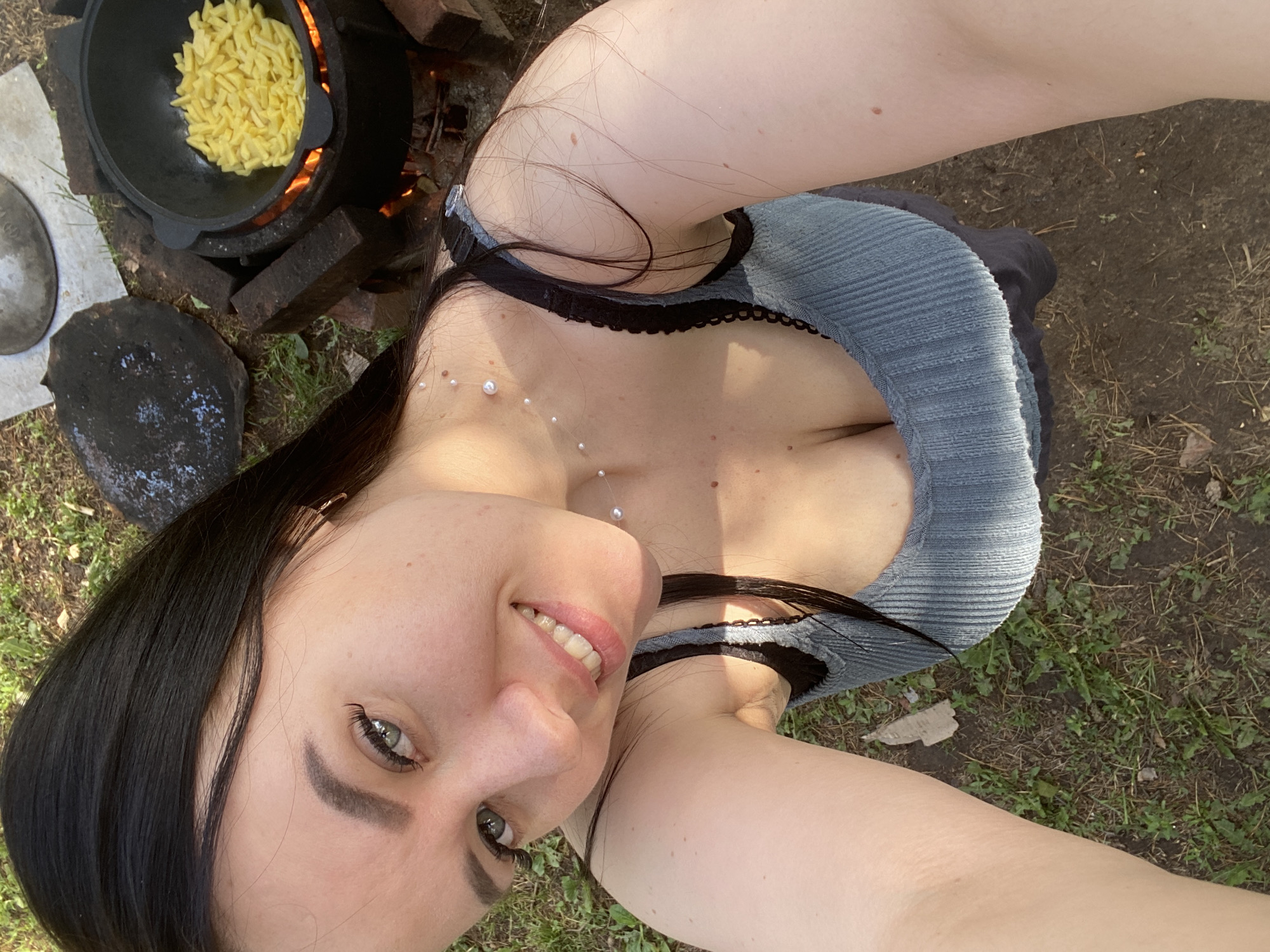 A woman is taking a selfie outdoors, likely near a campsite or cooking area. To her upper left, a black cast iron pot sits atop a makeshift brick stove with a fire burning inside. The pot contains yellow pasta or noodle-like food. The woman is fair-skinned with dark hair and is wearing a low-cut ribbed blue top with black lace detailing around the neckline. She is smiling at the camera. The ground around her is covered in grass and dirt. There are additional cooking utensils near the pot. The specific location within the city of Chaikovsky, Russia, cannot be determined from the image.