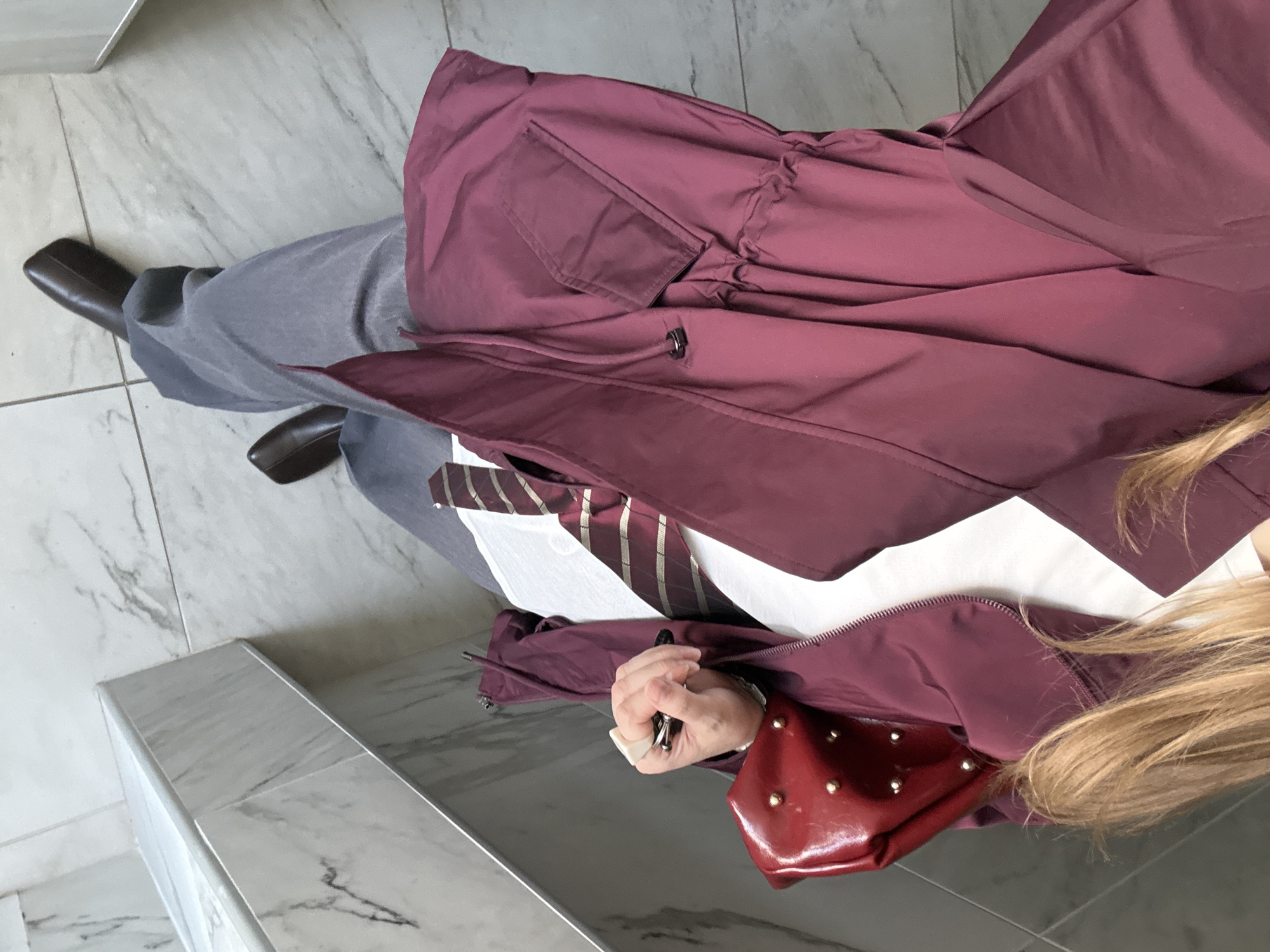 A person in a maroon coat and gray pants stands on a marble floor. They wear dark brown shoes and a striped tie. The person holds a pen in their hand, and a red purse is visible. The location appears to be indoors. There is no evidence to confirm that the location is in the city of Izhevsk, Russia.