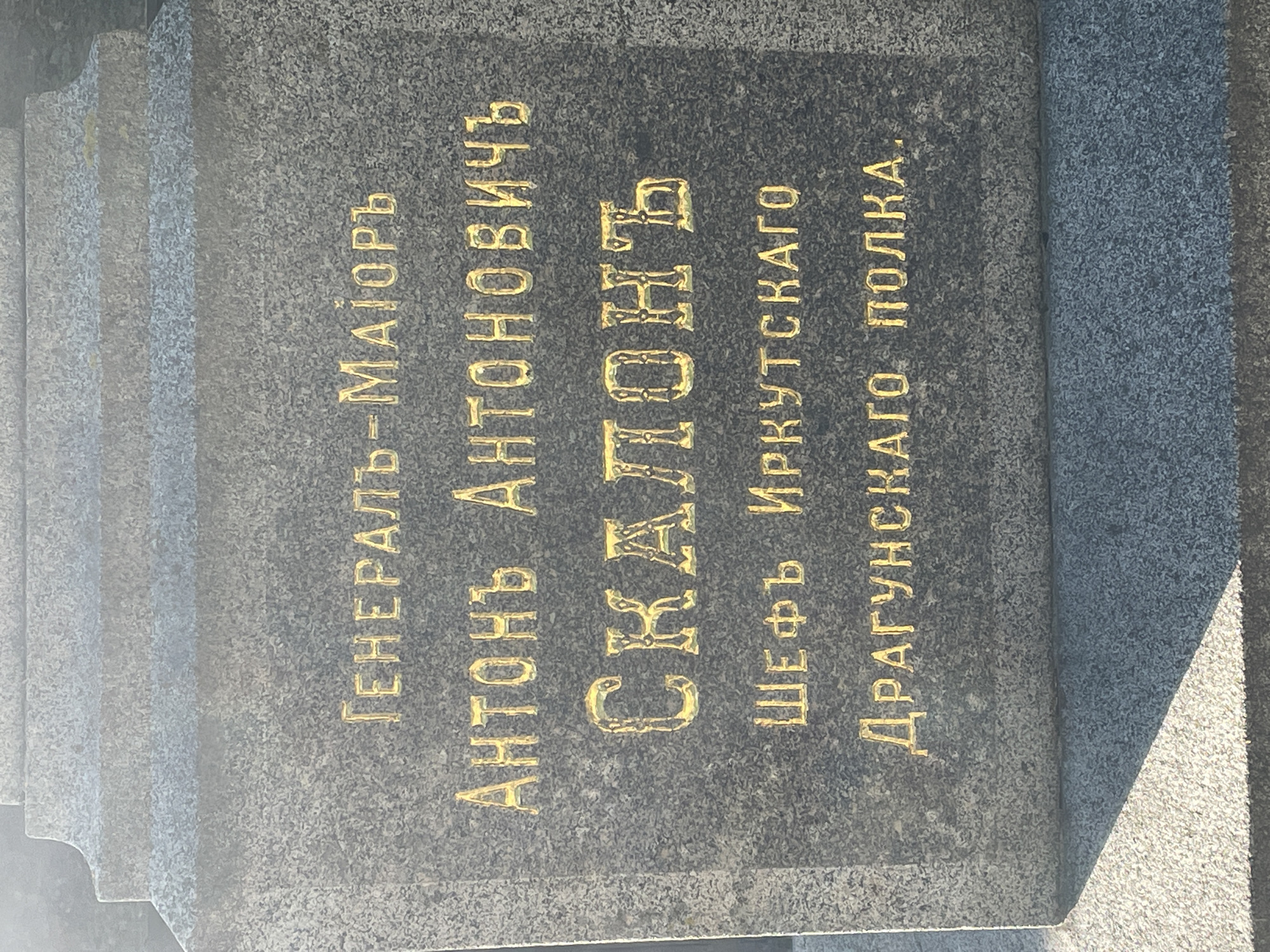The image shows a granite monument with gold lettering, likely a tombstone. The inscription is in Russian and appears to commemorate a military figure. The text reads: "ГЕНЕРАЛ-МАЙОРЪ АНТОНЪ АНТОНОВИЧЪ СКАЛОНЪ ШЕФЪ ИРКУТСКАГО ДРАГУНСКАГО ПОЛКА." This translates to "Major General Anton Antonovich Scalon, Chief of the Irkutsk Dragoon Regiment." The monument is set against a backdrop of gray, tiered stone, and a shadow suggests it is outdoors. The lighting indicates it is daytime, possibly sunny. There are no people or animals visible in the image. The location is identified as Sof'ino, Russia.