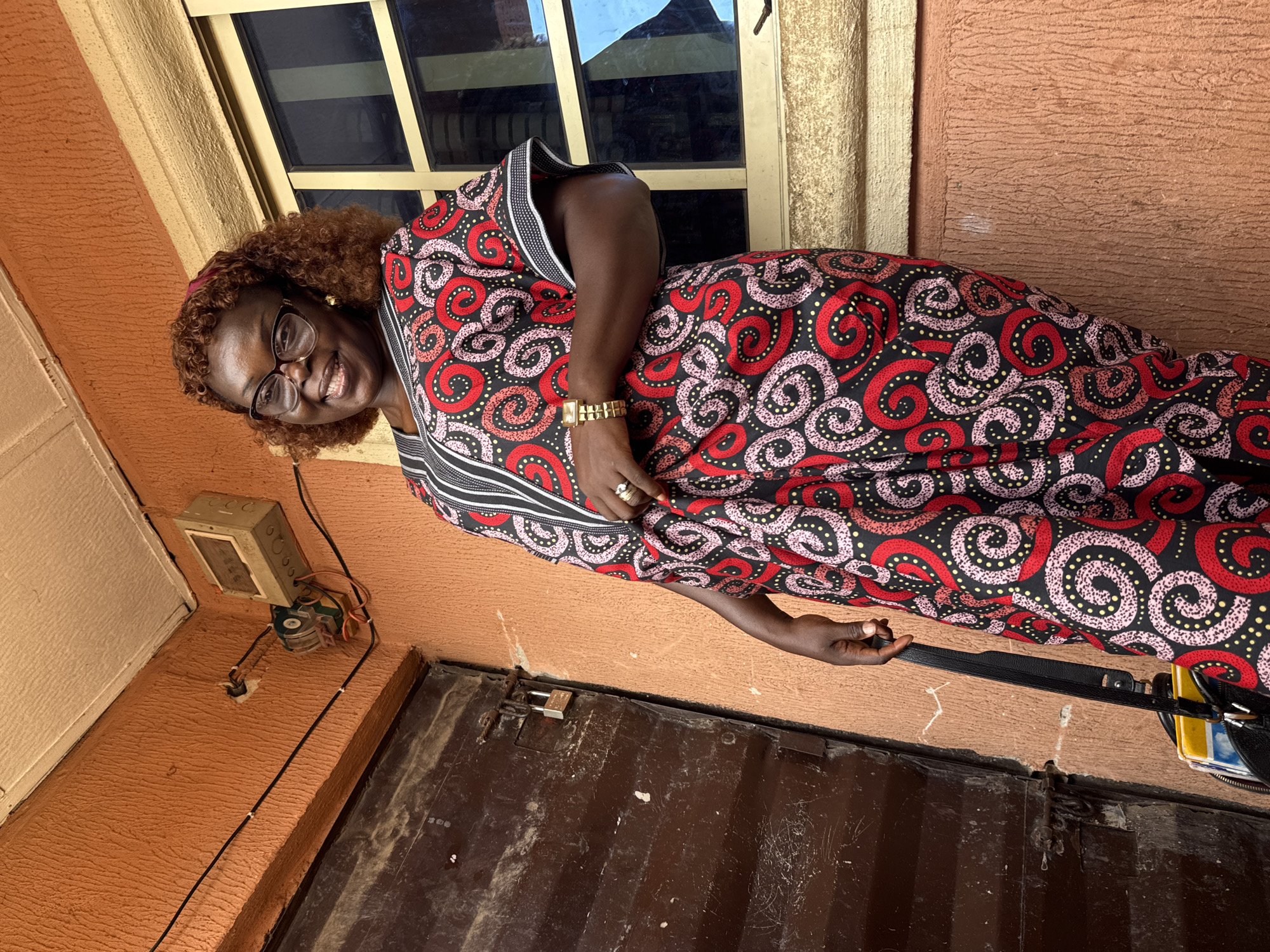 A middle-aged African woman is pictured standing outside a building. She is wearing a vibrant, patterned dress in red, pink, yellow, and black, along with eyeglasses. Her hair is styled in brown curls, and she wears a gold watch and a ring. She smiles towards the camera. The background includes a peach-colored wall, a window with bars, an electrical box with exposed wiring, and a brown metal door that appears locked. Based on the architectural style and clothing, the location is likely in the city of Onitsha, Nigeria. The picture shows the woman posing in front of the building with a purse strap in her hand.