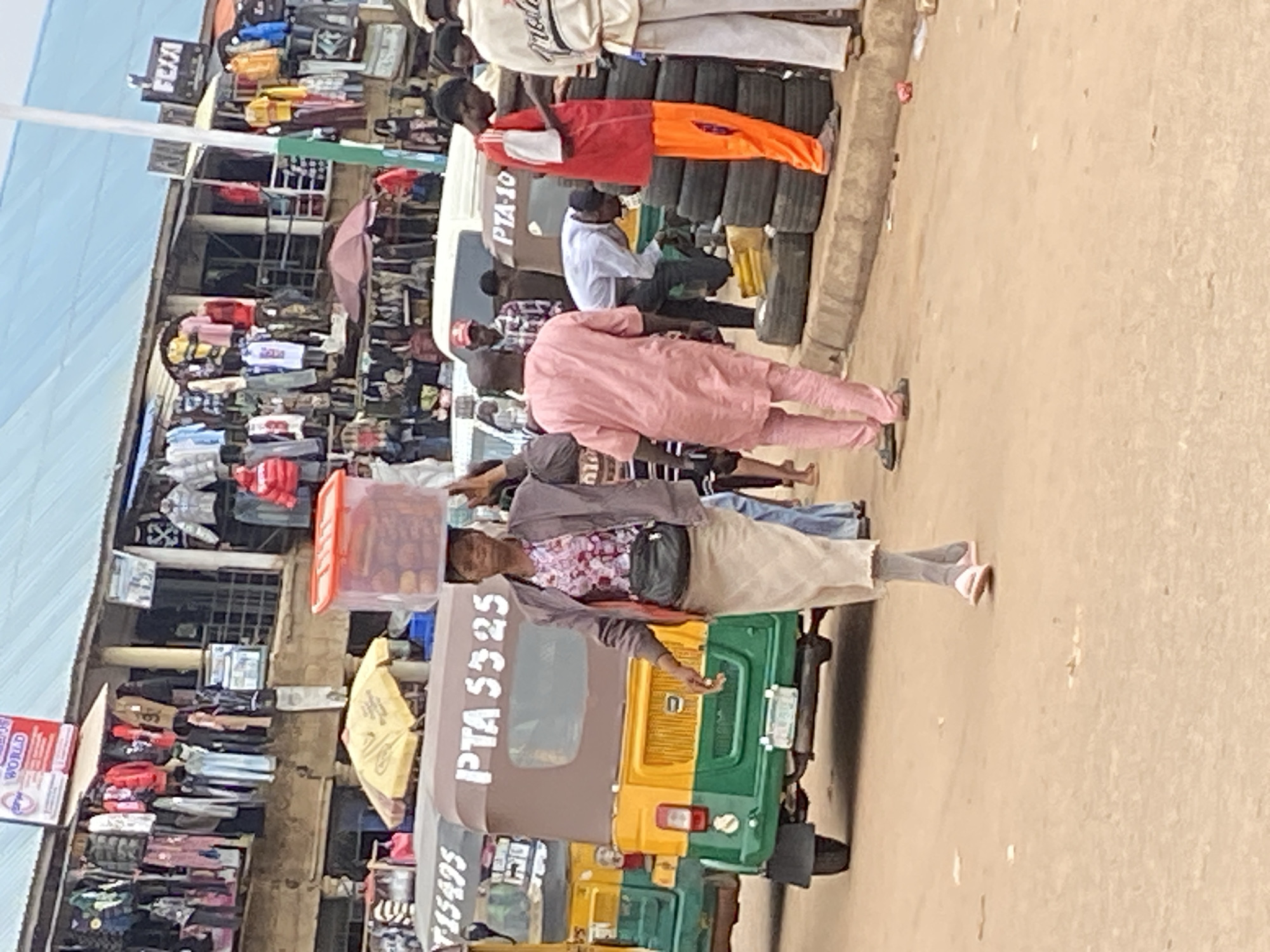 A medium shot captures a street market scene in Bukuru, Nigeria. In the foreground, a woman walks from left to right, carrying a clear plastic container filled with brown items on her head. She wears a patterned short-sleeved shirt, a light-colored skirt, gray socks, and white slip-on shoes. A black crossbody bag is slung over her right shoulder. To her left, a green and yellow motorized tricycle with the inscription "PTA 5325" is parked, facing away from the viewer. Behind the tricycle, another similar tricycle, partially visible, has "15495" written on its side.
The background reveals stalls lining the street, displaying various clothing items. These stalls are set up under a corrugated metal roof. In the mid-ground, several people are present. A man in a pink kaftan walks away from the camera. To his right, a man in a white shirt and dark pants is bent over, appearing to work on something near a stack of black tires. Another man in a red shirt and orange pants stands near the tires, looking at his phone. Further back, more people are visible amidst the stalls and parked vehicles, indicating a busy marketplace. The ground is sandy and dusty. The overall atmosphere suggests a typical day in a bustling Nigerian market.
