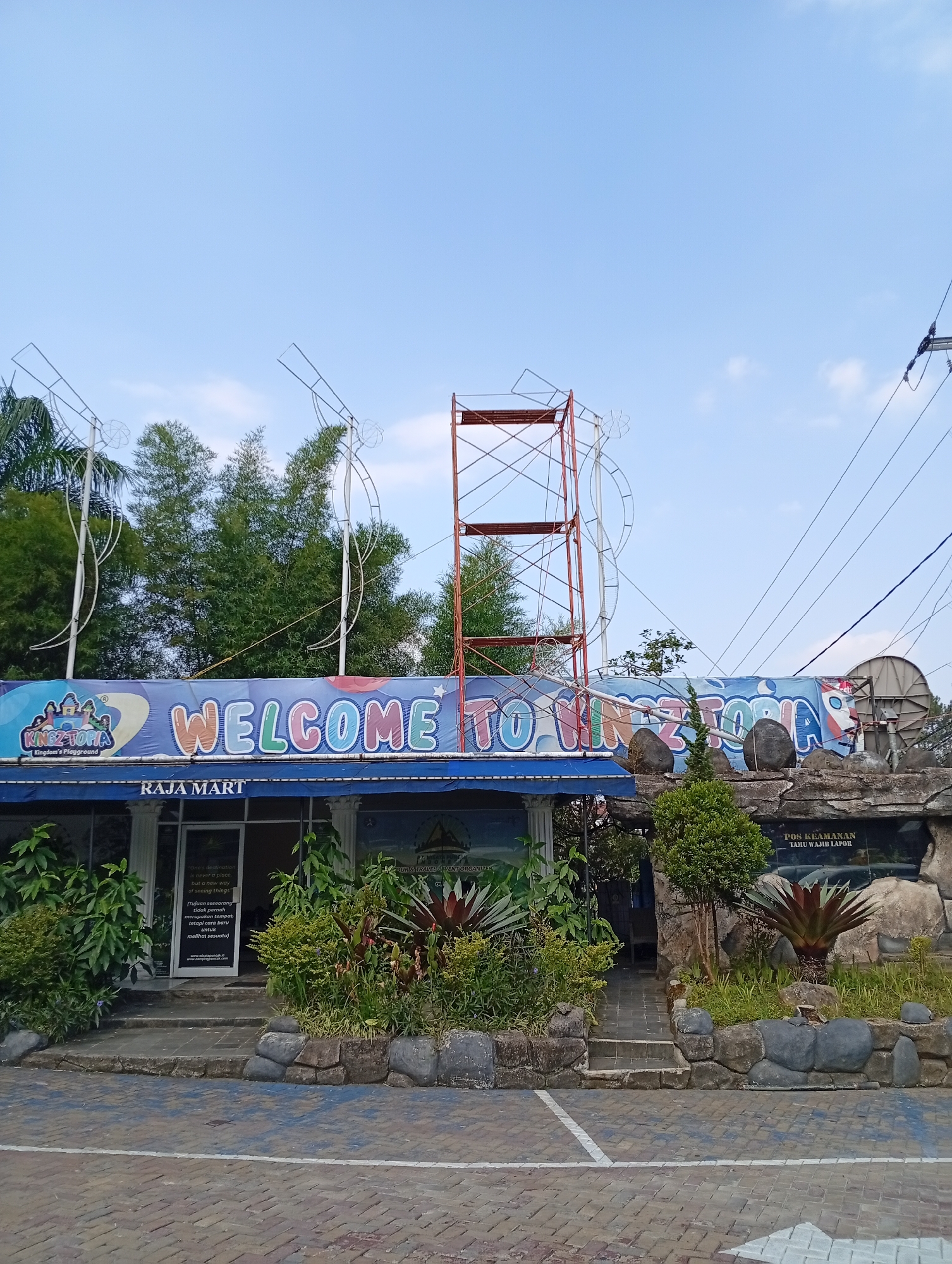 The image shows the entrance to a facility identified by a large banner that reads "WELCOME TO KINGZ-TOPIA Kingdom's Playground." Below this banner, another sign indicates "RAJA MART." The entrance appears to be a shop or service area with glass doors and windows, surrounded by landscaping featuring various plants and large stones. To the right of the entrance, there is a rocky facade with signage that says "POS KEAMANAN TAMU WAJIB LAPOR" (Security Post, Guests Must Report). Overhead, there are metal structures and wires, suggesting some ongoing construction or part of the attraction's setup. The overall scene is outdoors under a clear blue sky, with trees and bamboo visible in the background. The location is identified as Cimacan, Indonesia.
