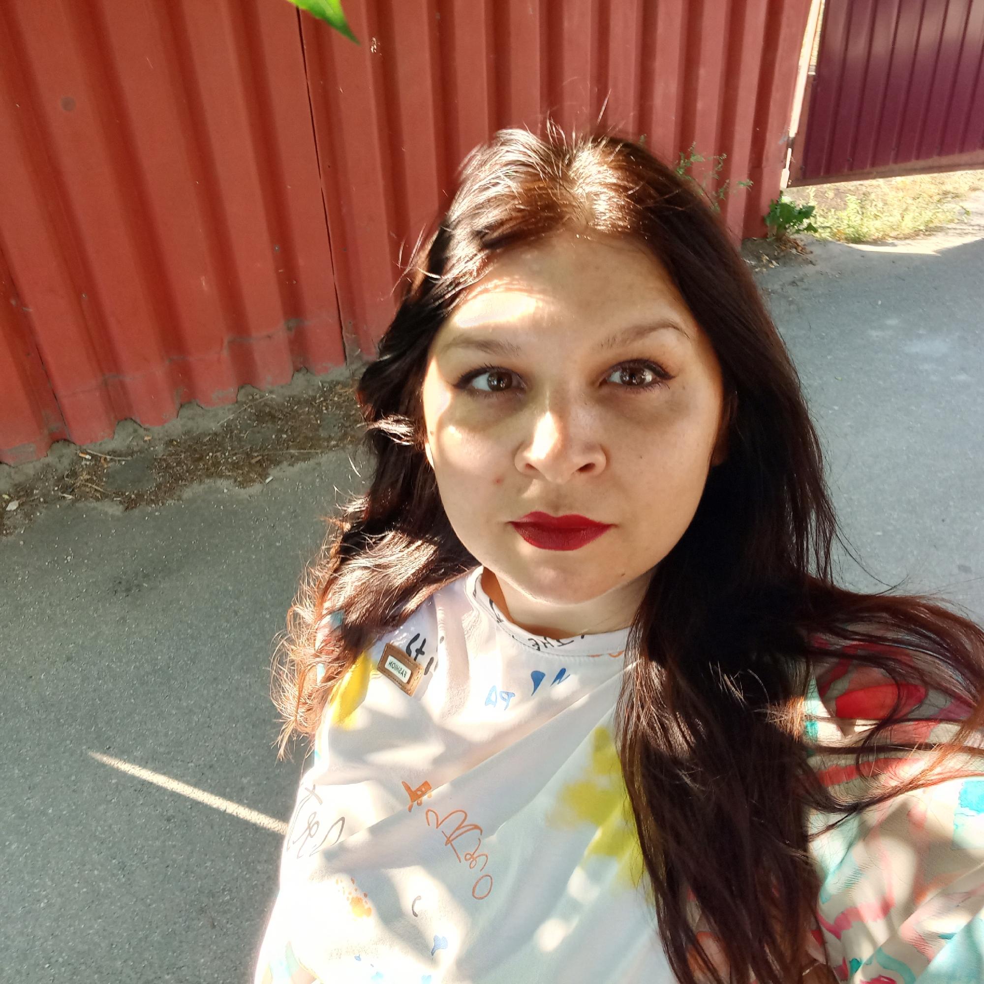 A woman with long, dark brown hair and brown eyes takes a selfie outdoors. She is wearing a white t-shirt with colorful abstract designs and text. Her lips are painted a deep red. Sunlight casts shadows across her face, highlighting one side. Behind her, a corrugated metal fence, painted a faded red, runs vertically. To the right of the fence, a darker, maroon-colored gate is visible, slightly ajar. The ground is paved with asphalt, and there are patches of dry grass and weeds near the fence. The scene appears to be an outdoor area, possibly a residential yard or alleyway, located in Kropyvnytskyi, Ukraine, given the style of the fence and the general environment.