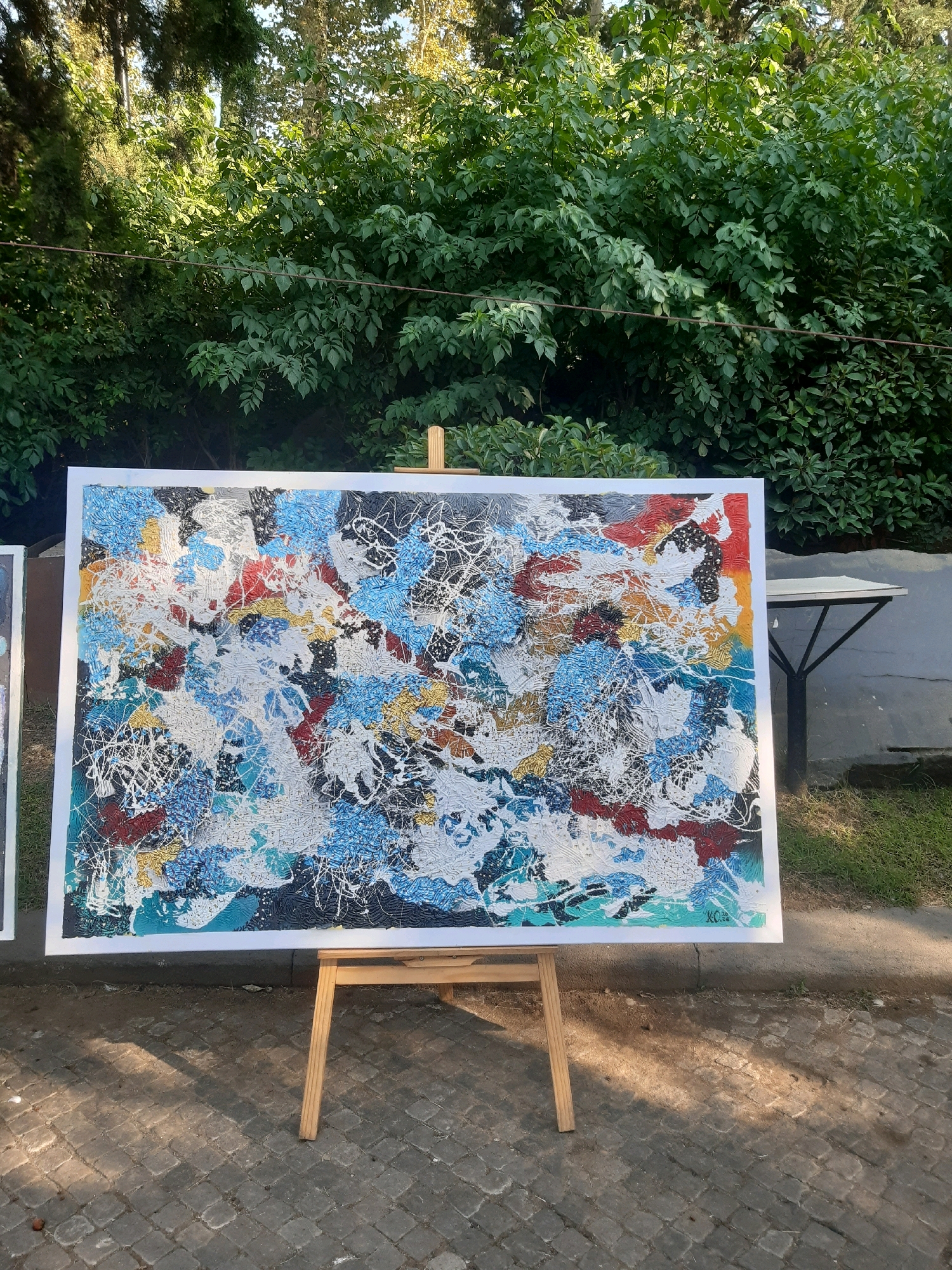 An abstract painting on a white canvas is displayed outdoors on a wooden easel. The painting is vibrant and textured, with a chaotic mix of colors including blues, whites, reds, blacks, and gold. Splattered and layered paint creates a dynamic surface. Behind the painting, lush green foliage from trees and bushes dominates the background. Sunlight filters through the leaves, casting dappled shadows on the cobblestone ground in front of the easel. To the right of the painting, a metal table with a gray surface and black legs is visible. The scene appears to be in a city park or an outdoor exhibition area, possibly in Tbilisi, Georgia, given the context of other images provided. No people are present in the frame.