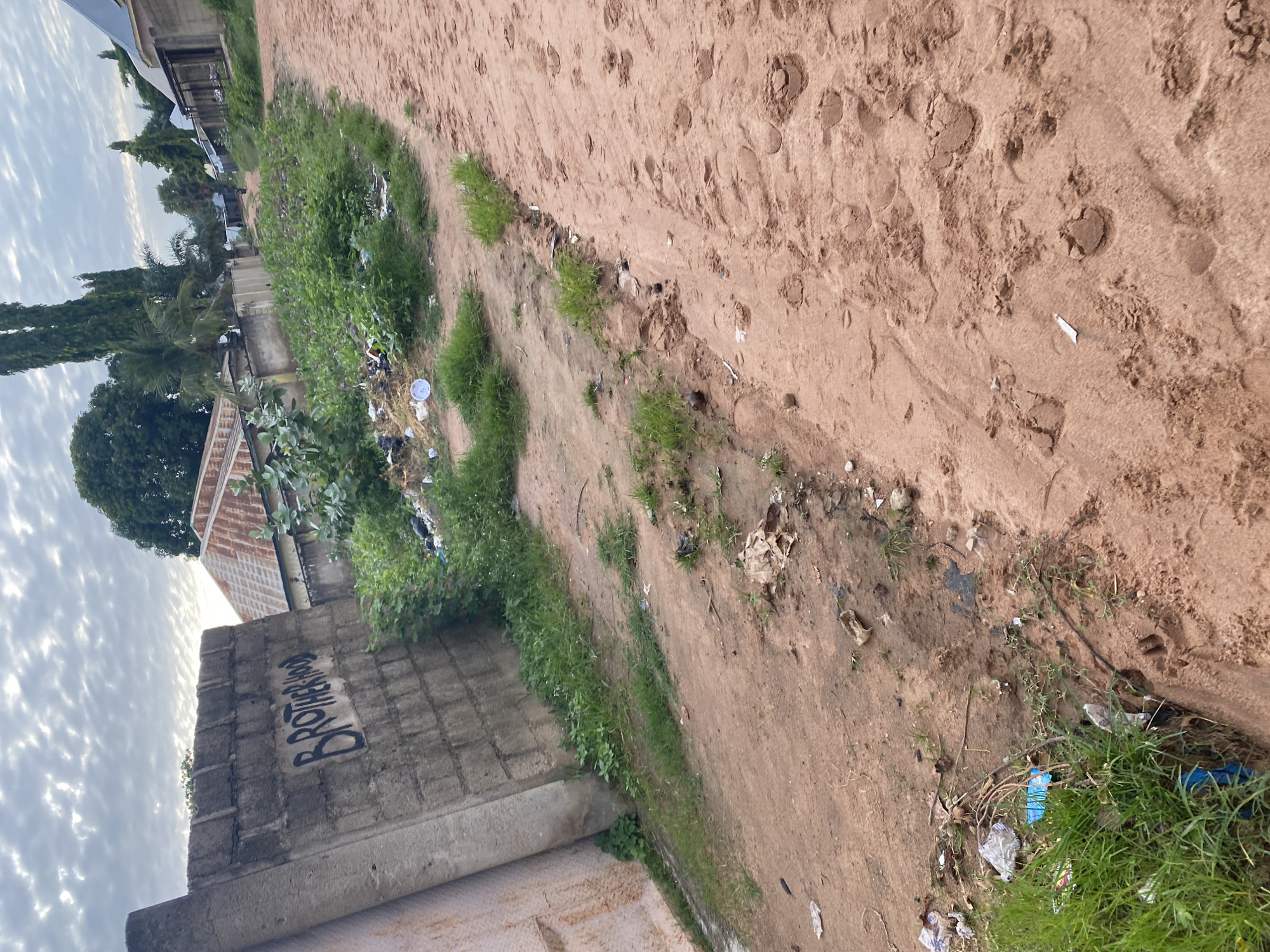The image depicts a dirt road in Jauro Yinu, Nigeria, with a concrete wall on the left side. The wall is graffitied with the word "BROTHERHOOD" in black spray paint. The road is unpaved and appears to be a rural or semi-rural area, with vegetation and scattered debris along its edges. The sky is overcast with clouds. Buildings and trees are visible in the background, suggesting a residential area. There are footprints in the dirt road, indicating recent passage of people or animals. The overall scene suggests a somewhat neglected environment with evidence of human activity.