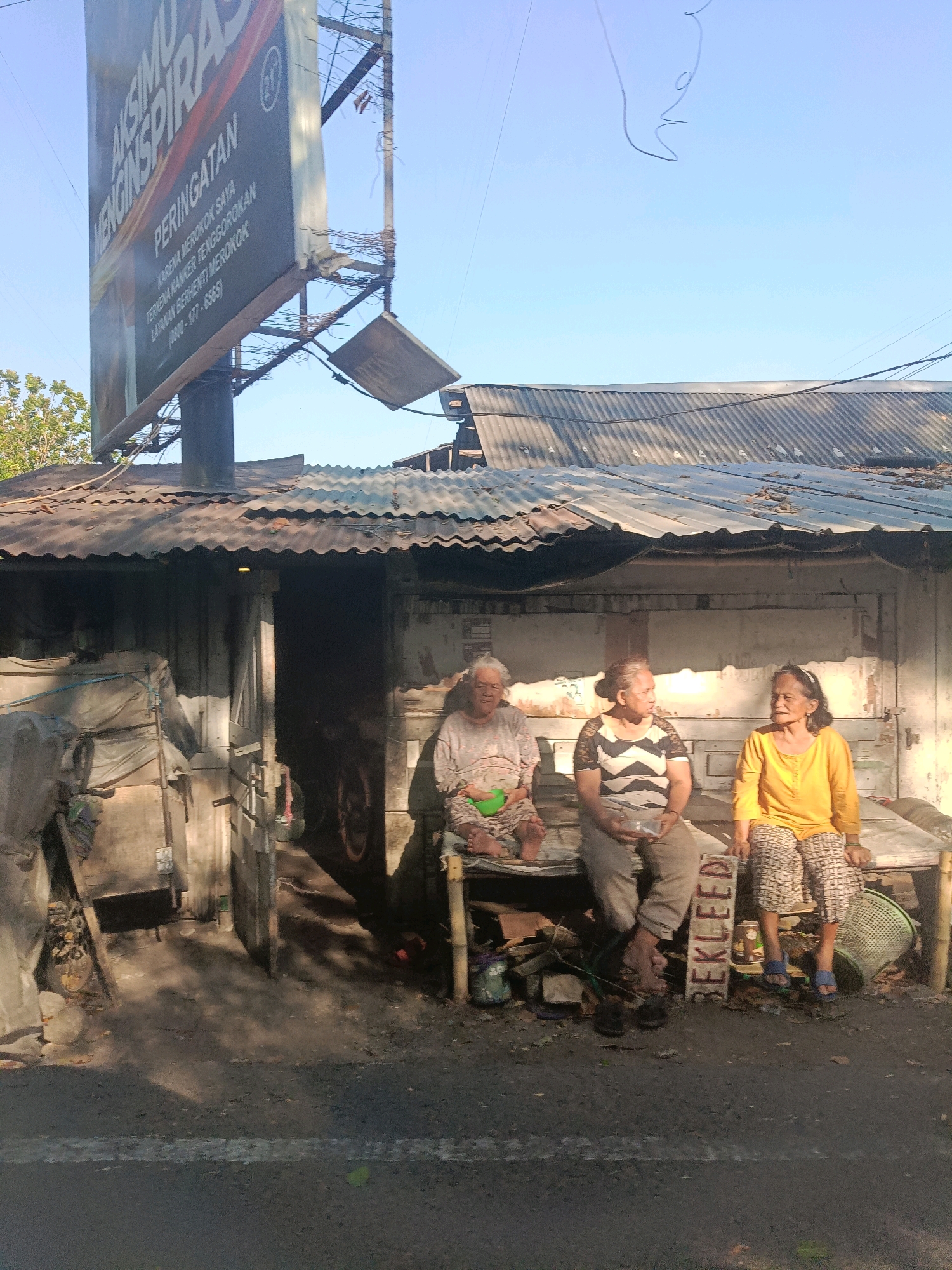 The image depicts a street scene in Surabaya, Indonesia, with three elderly women sitting on a wooden bench outside a weathered structure. The building has a corrugated metal roof and wooden walls. The woman on the left wears a light-colored patterned top and pants, holding a green bowl. The woman in the middle is dressed in a black and white striped shirt and grey pants, holding a small white object. The woman on the right wears a bright yellow long-sleeved shirt and patterned pants, with blue sandals. In front of them, there is a collection of objects including a woven basket, stacks of wood, and various containers. A faded billboard advertising "AKSIMU MENGINSPIRASI" and a health warning regarding smoking is visible in the background, attached to a damaged metal pole. The ground is asphalt with some debris. The lighting suggests it is daytime, with strong shadows cast by the structures and objects.