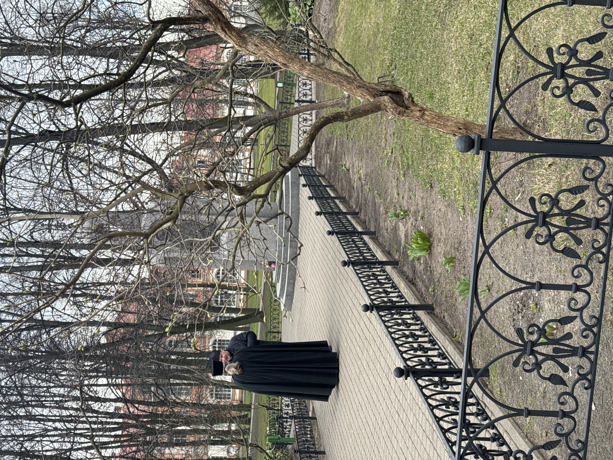 The image depicts a person dressed in a black religious robe and headwear, possibly a cassock, standing on a paved walkway in an outdoor setting. The walkway is bordered by a decorative black iron fence on one side and a patch of ground with grass and sparse vegetation on the other. In the background, bare trees with budding branches are visible, partially obscuring the view of a building with multiple windows. Based on the architectural style and the presence of religious attire, it is plausible that the location is within the city of Saint Petersburg, Russia, near a religious building. The person is facing the viewer.