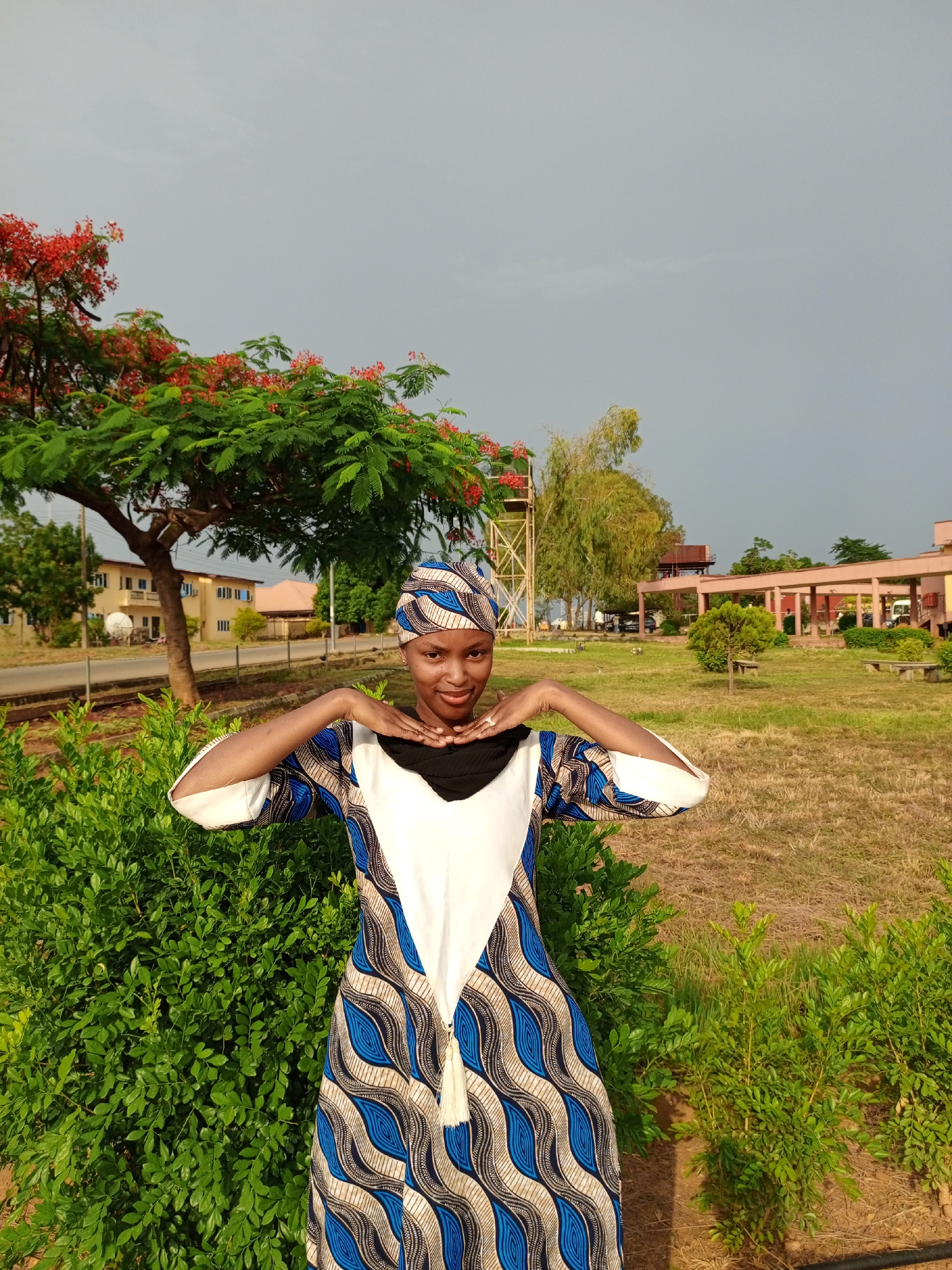 A woman stands outdoors in Keffi, Nigeria, framed by greenery. She wears a patterned headwrap and a dress with a white bodice and blue, black, and tan patterned sleeves and skirt. Her hands are raised, framing her face. Behind her, there is a manicured lawn with trees, buildings, and a water tower in the background. The sky is overcast.