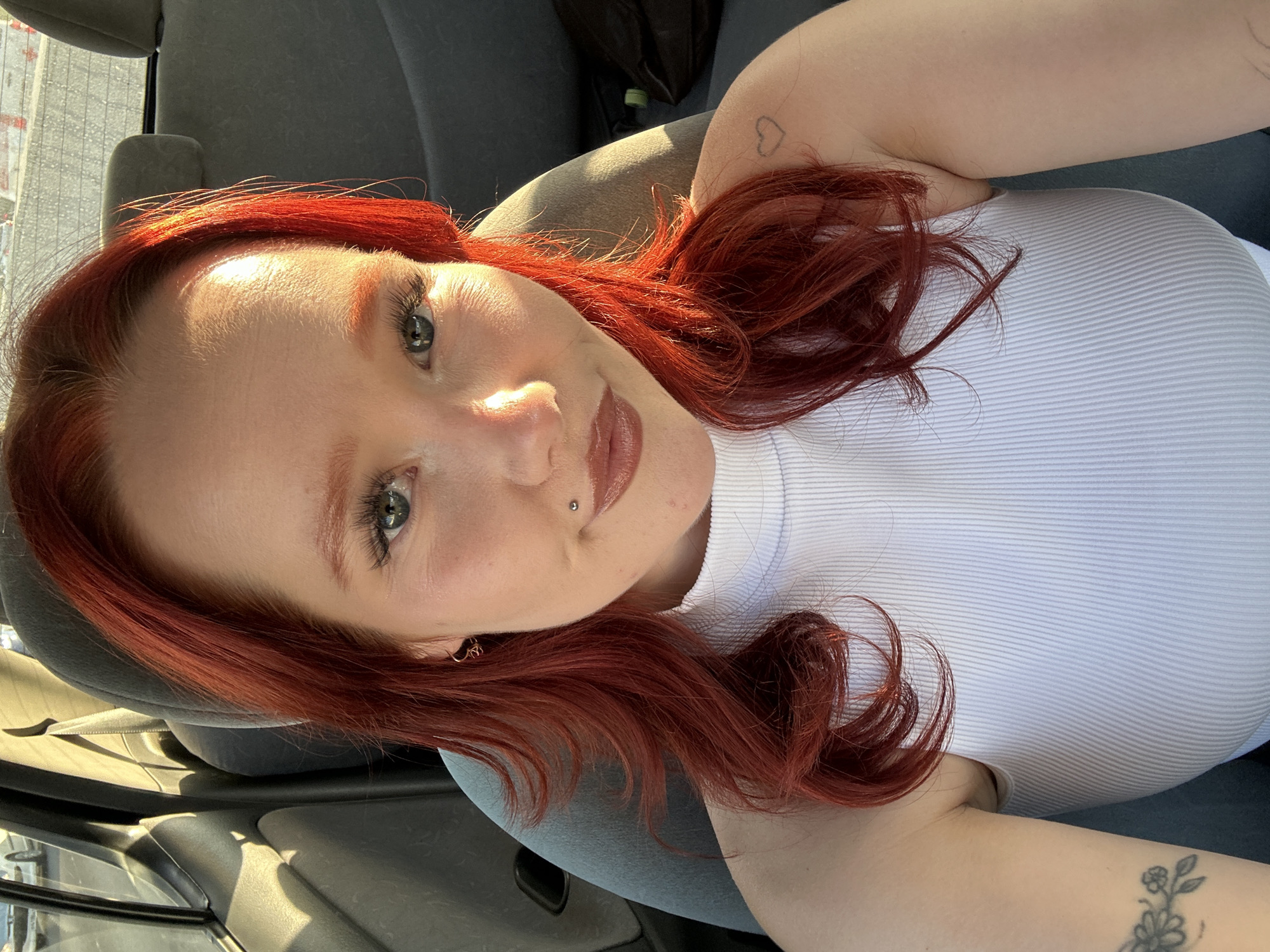 The image shows a young woman with vibrant red hair in a selfie taken inside a car. She is wearing a white ribbed tank top. Her hair is styled in loose waves, with some strands falling around her face. She has a piercing on her lower lip and a small heart tattoo on her upper arm. The sunlight is casting strong shadows, suggesting it is daytime. The background is blurred, but appears to be the interior of a vehicle with gray upholstery. The woman's expression is relaxed and her eyes are looking directly at the camera. There is no visible text in the image.