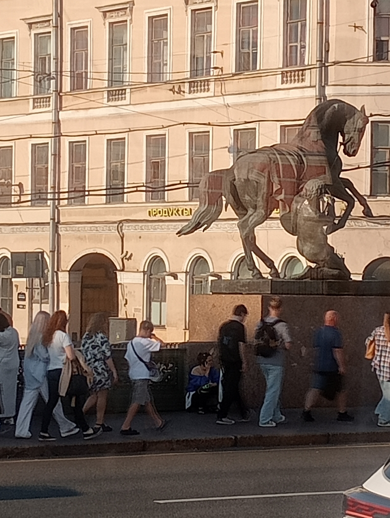 The image shows a street scene in Saint Petersburg, Russia. In the background, a building with classical architecture and many windows is visible. Above an arched entrance, the word "ПРОДУКТЫ" (Products) is written in yellow letters. In the foreground, a bronze sculpture of a horse and a kneeling figure stands on a pedestal. The sculpture is ornate and detailed. Several people are walking along the sidewalk in front of the building and the sculpture. The group consists of men and women of various ages, dressed in casual clothing. One person is sitting on the ground near the sculpture, looking at something in their hands. The sun is shining, casting shadows, suggesting it is daytime. The scene appears to be a typical urban environment, possibly a tourist area given the presence of the prominent sculpture.