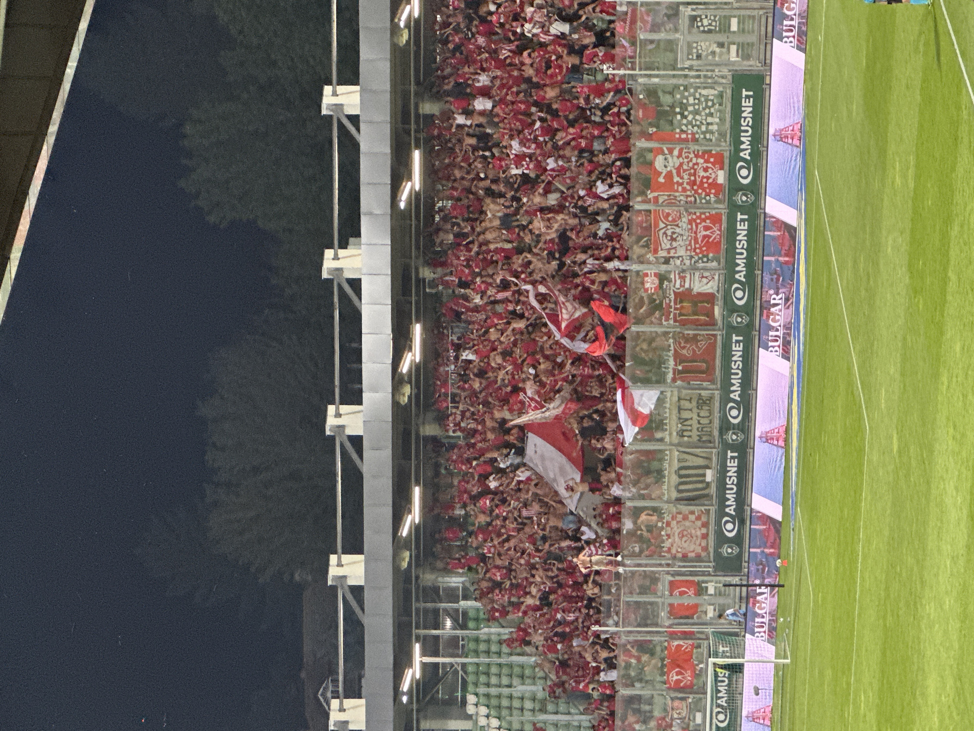The image shows a packed stadium filled with football fans wearing red. The fans are standing and appear to be cheering, with many holding flags and banners. The stadium is modern with tiered seating and floodlights. In the foreground, there is a green football pitch with advertising hoardings visible. One hoarding features the text "BULGAR". The scene is likely at the Huvepharma Arena, the home stadium of PFC Ludogorets Razgrad, located in the city of Razgrad, Bulgaria. The atmosphere suggests a live sporting event, possibly a football match, with a high level of fan engagement.