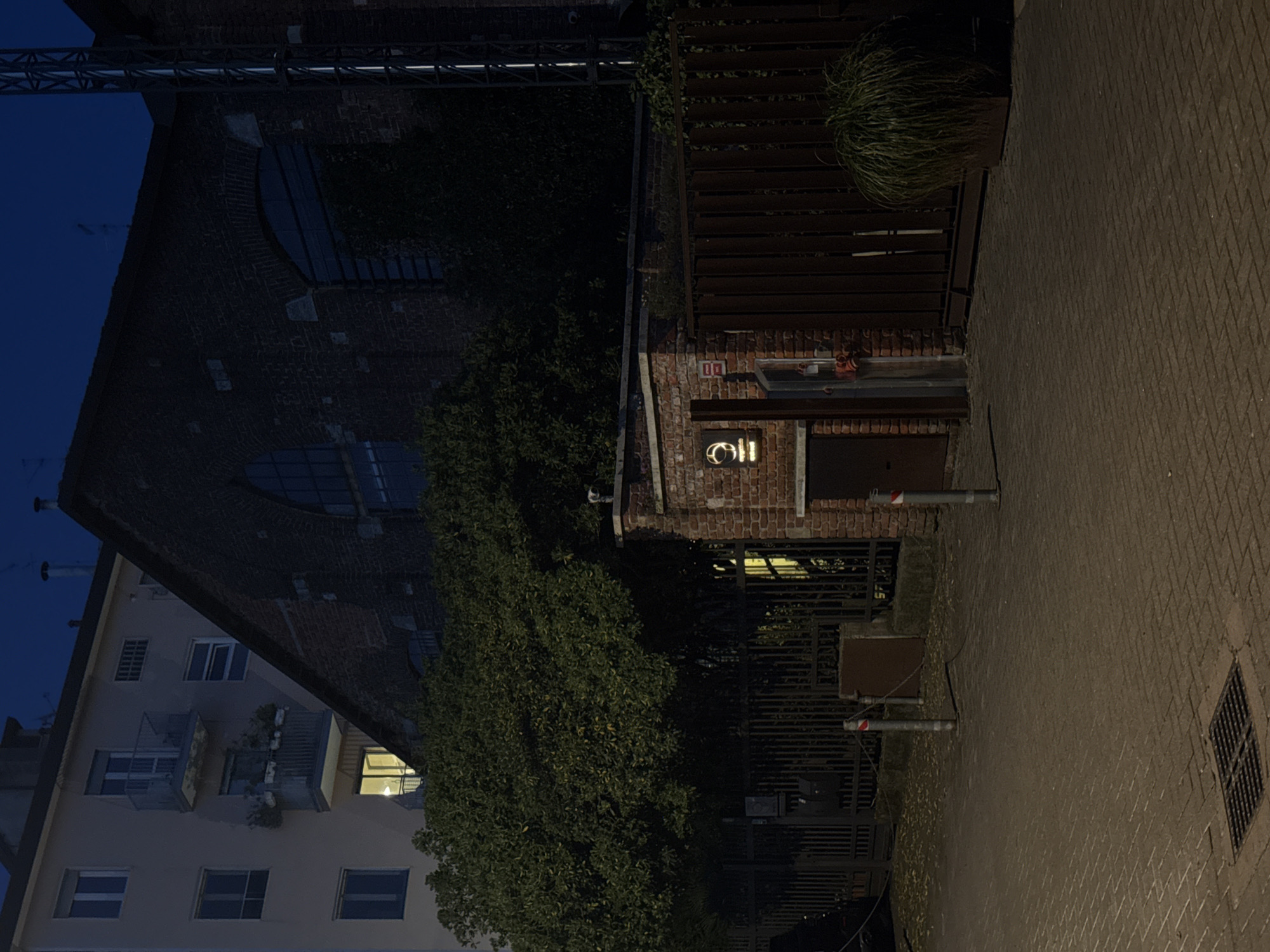 The image captures a nighttime scene in Milan, Italy, featuring a brick building with a sign that reads "OII" illuminated on its facade. A dark metal structure, possibly a trellis or fence, is visible atop the building. A large bush or tree obscures part of the foreground and the building's lower section. The scene transitions to a paved walkway or street on the right, showing brick patterns and a metal grate. The adjacent building on the left displays residential architecture with lit windows and balconies. The sky is dark, suggesting a late evening or early morning setting.