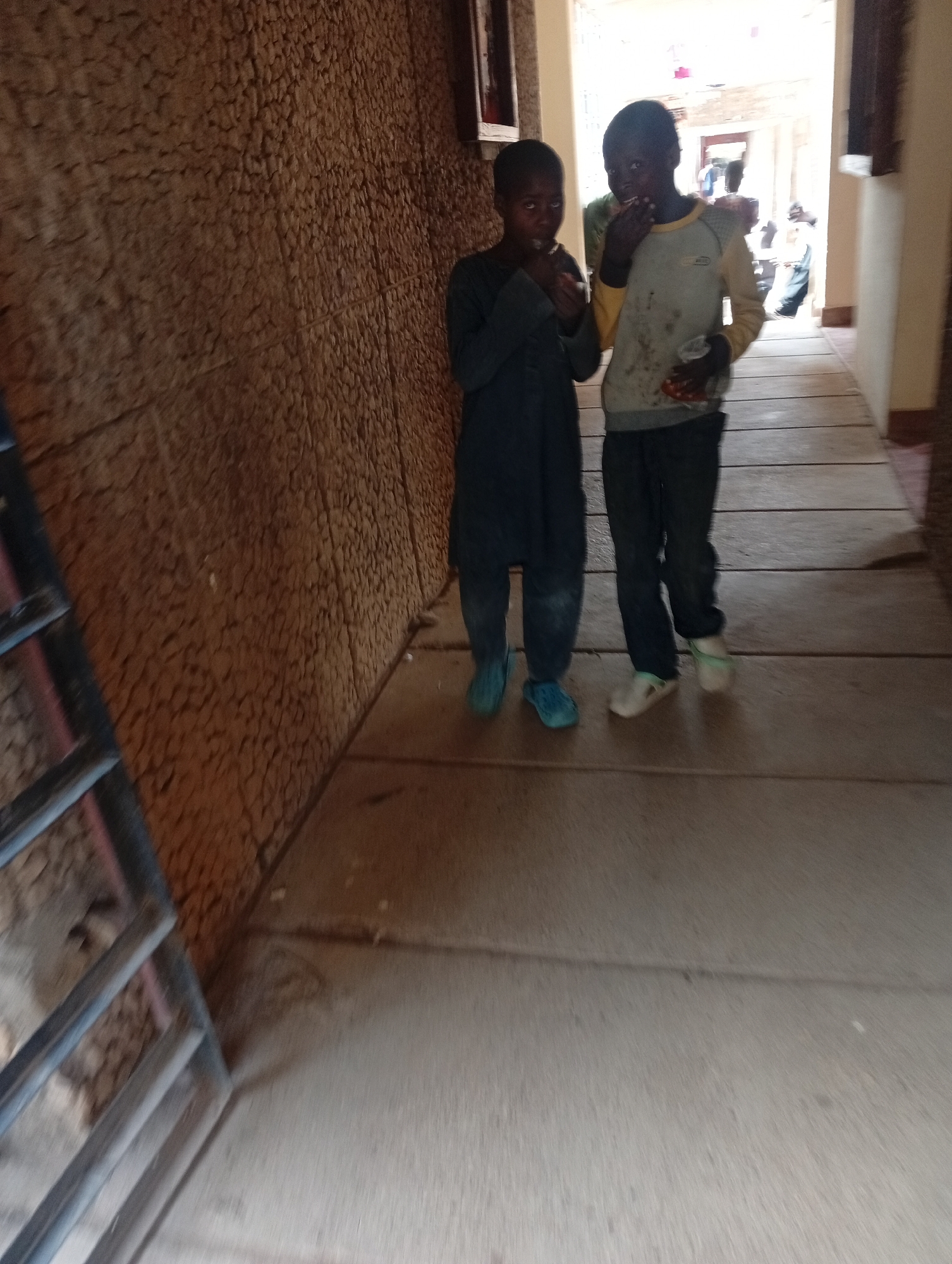 In a dimly lit hallway with a rough, textured brown wall on the left, two young boys are walking towards the viewer. The boy on the left wears a dark, long-sleeved garment and light blue slip-on shoes. He appears to be eating something from his right hand. The boy on the right wears a light yellow and grey long-sleeved shirt, dark pants, and white slip-on shoes with a green detail. He holds a clear plastic cup in his left hand and is also eating something. His right hand is near his mouth. In the background, further down the hallway, other figures and bright light suggest an exit or opening. The floor is made of wooden planks. A metal railing is visible on the far left of the frame.