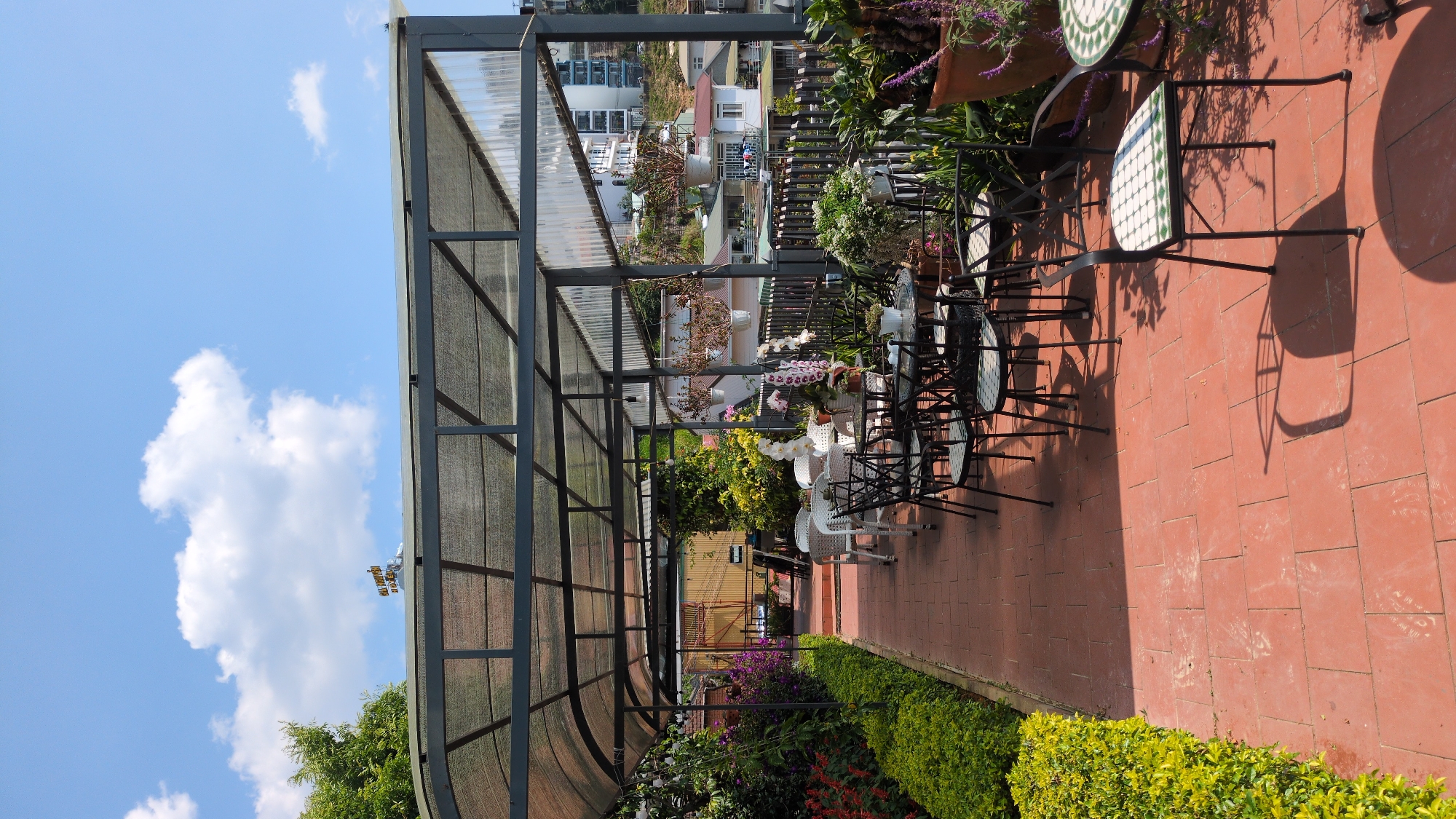 The image shows a view on a patio or outdoor seating area. The location is likely in Da Lat, Vietnam, based on the architectural style and common landscaping. The sky is blue with scattered clouds, and a partially transparent, metal-framed structure provides shade. There are tables and chairs with tile designs. Along the patio's edge, there's an arrangement of flowers and greenery. In the background, there are building roofs and foliage typical of a cityscape nestled in a hilly or mountainous area.