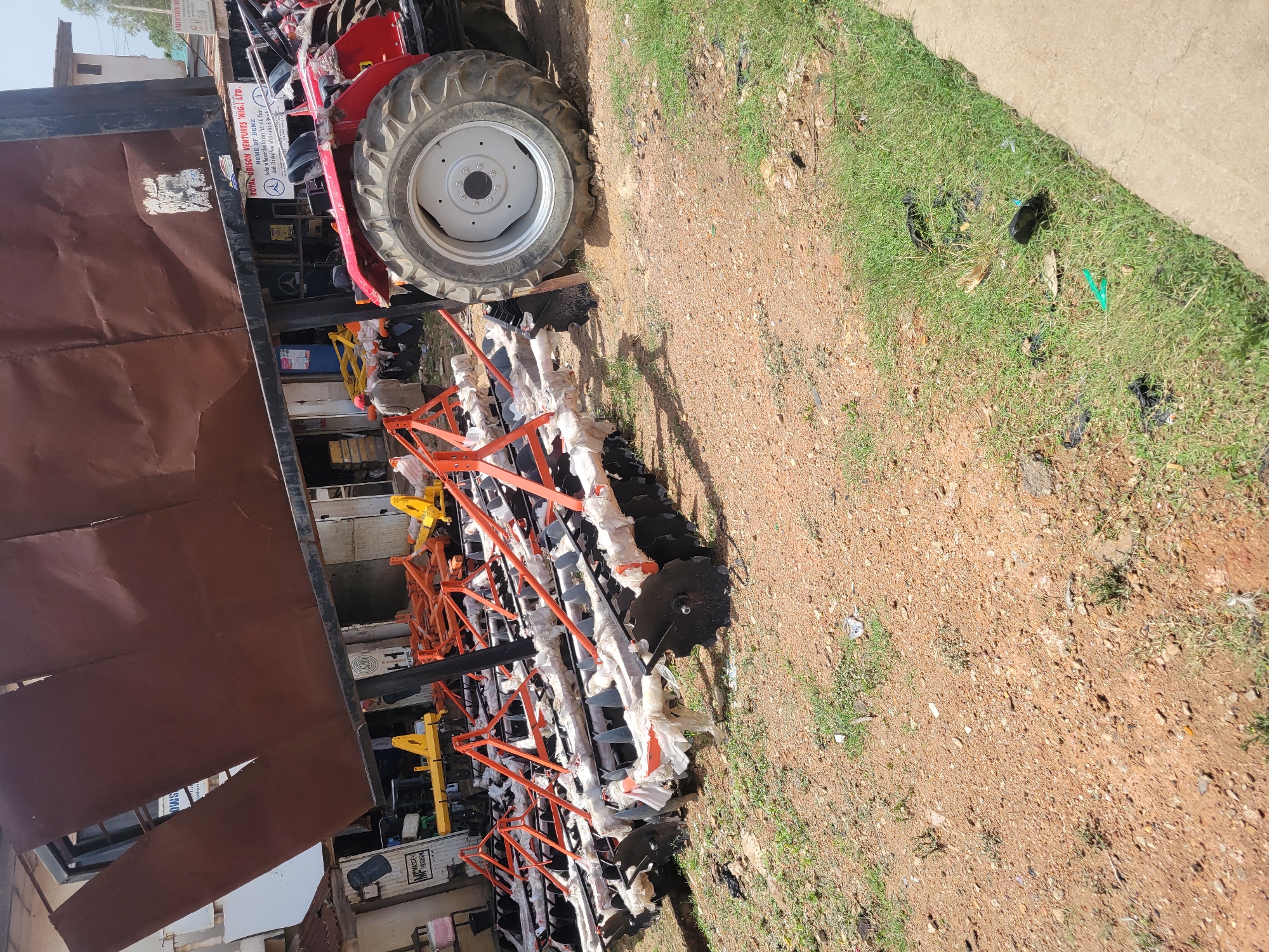 The image depicts a tractor parked on a dirt and grass-covered area, likely in a commercial or industrial setting. Attached to the tractor is a collection of agricultural implements, possibly disc harrows, wrapped in white plastic and secured with orange frames. The presence of a tractor and agricultural machinery suggests this location could be related to farming or equipment sales. The location is in the city of Unknown, Unknown.
---